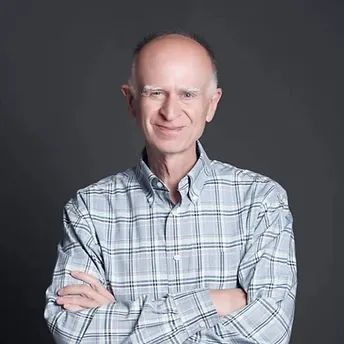 Bald man in a blue and white plaid shirt with arms crossed, smiling, against a gray background.