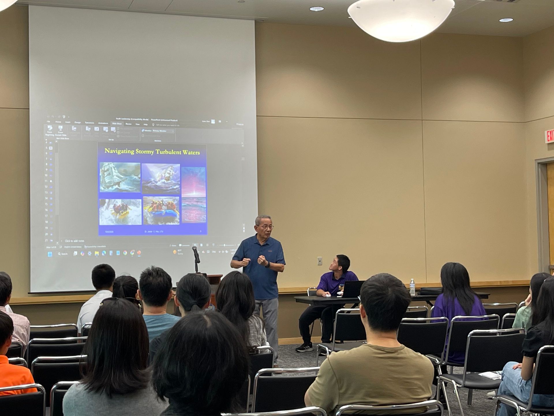 A man lectures in front of a screen to a seated audience in a brightly lit room.