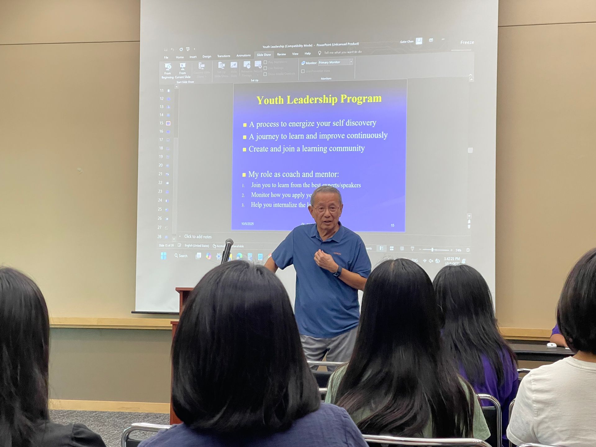 Man lecturing in front of an audience, a projected blue screen with text behind him.