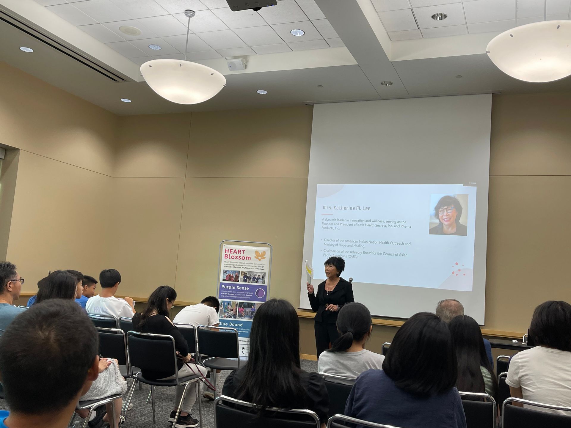 A woman gives a presentation in a room with a screen. Audience members sit and listen.