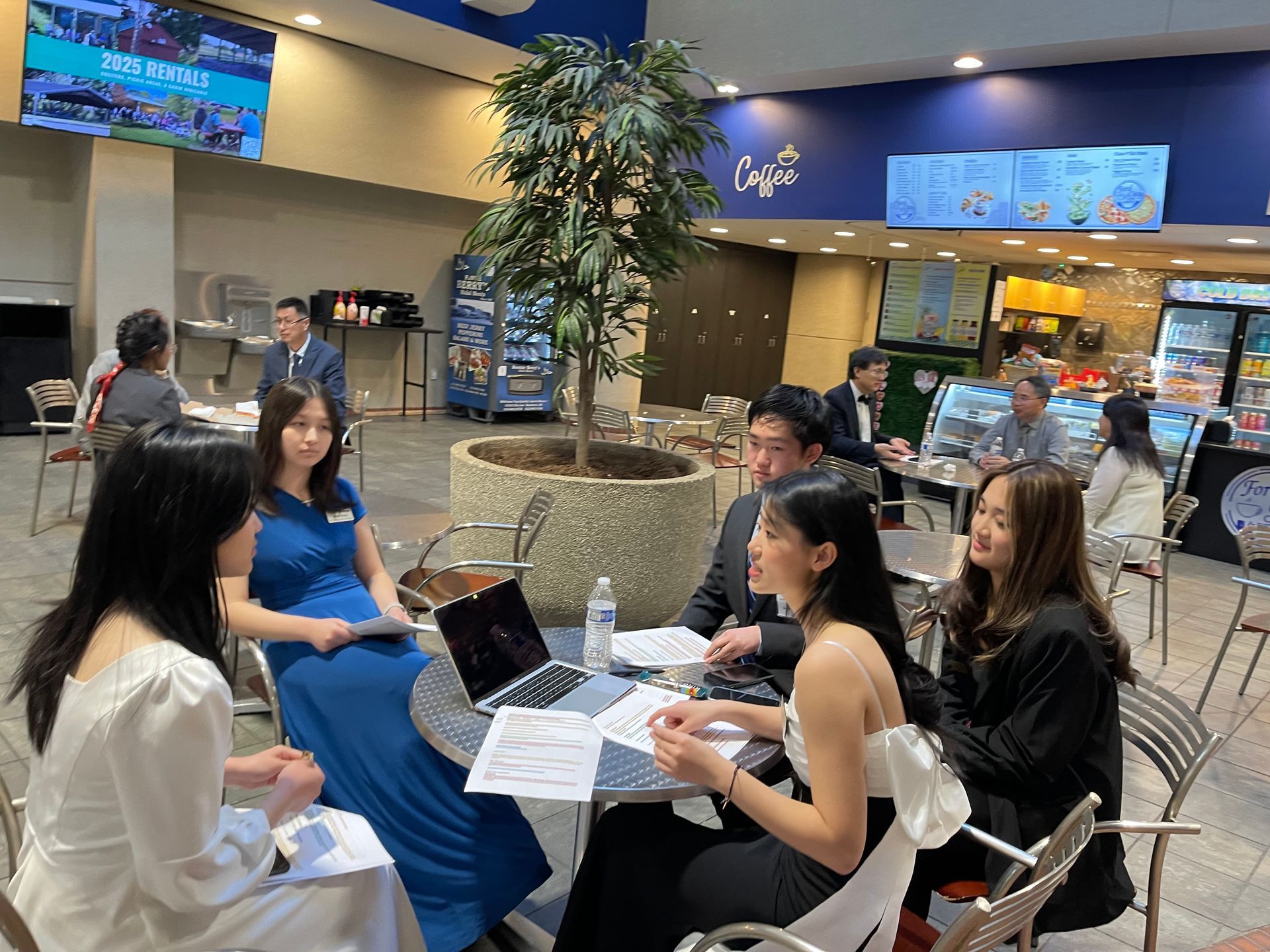 Group of young adults in formal attire at a cafe table, discussing documents near a laptop.