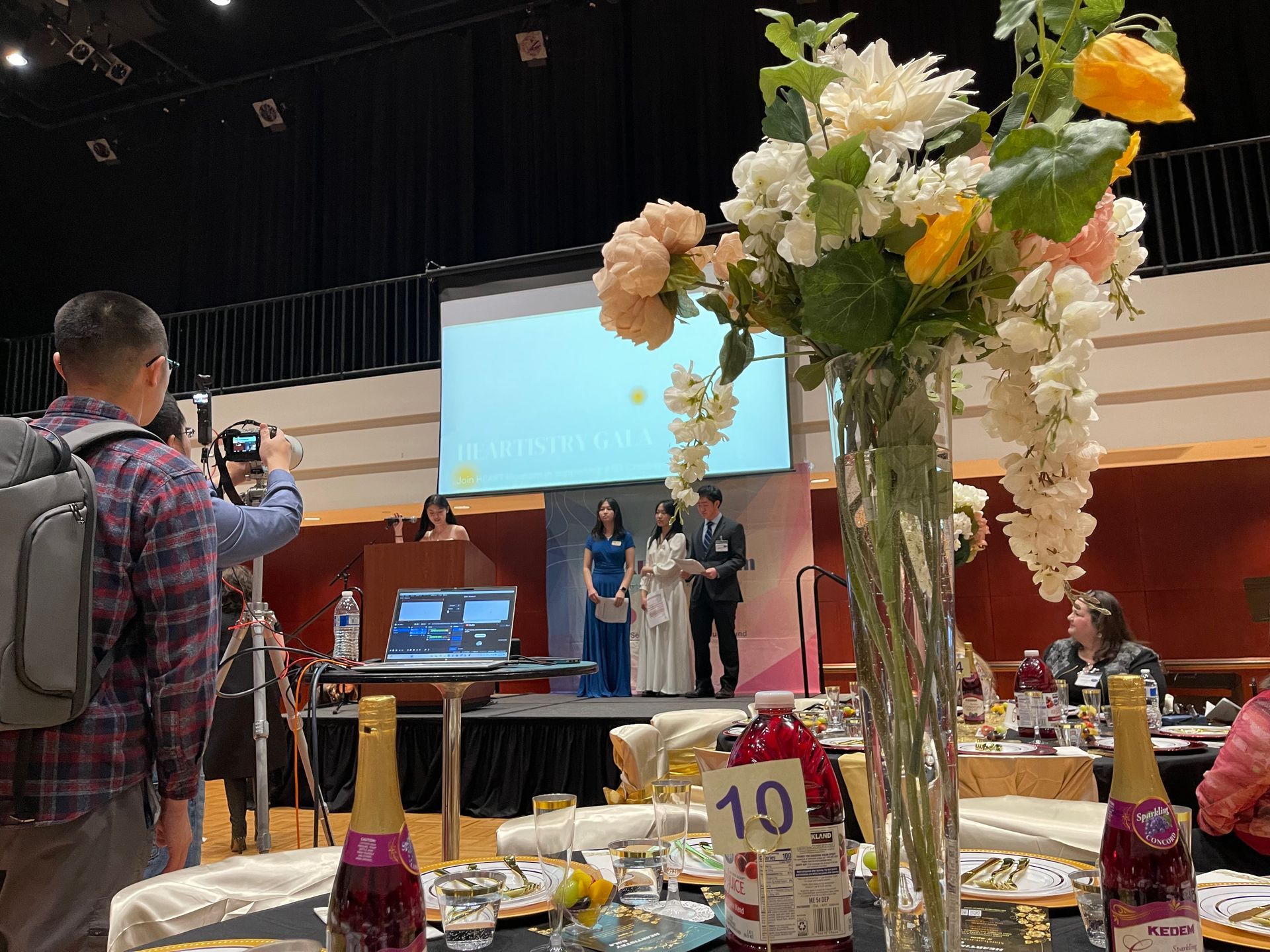 Formal event: Speakers on stage, flowers, photographer, table setting, audience.