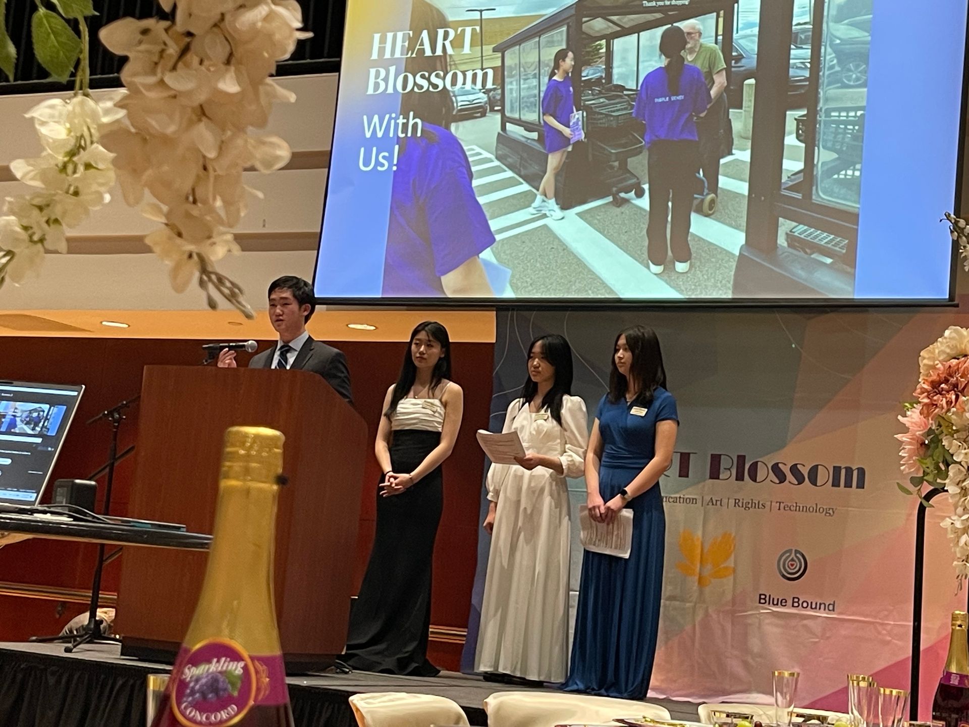 Man speaking at podium, three women beside him, screen displays 