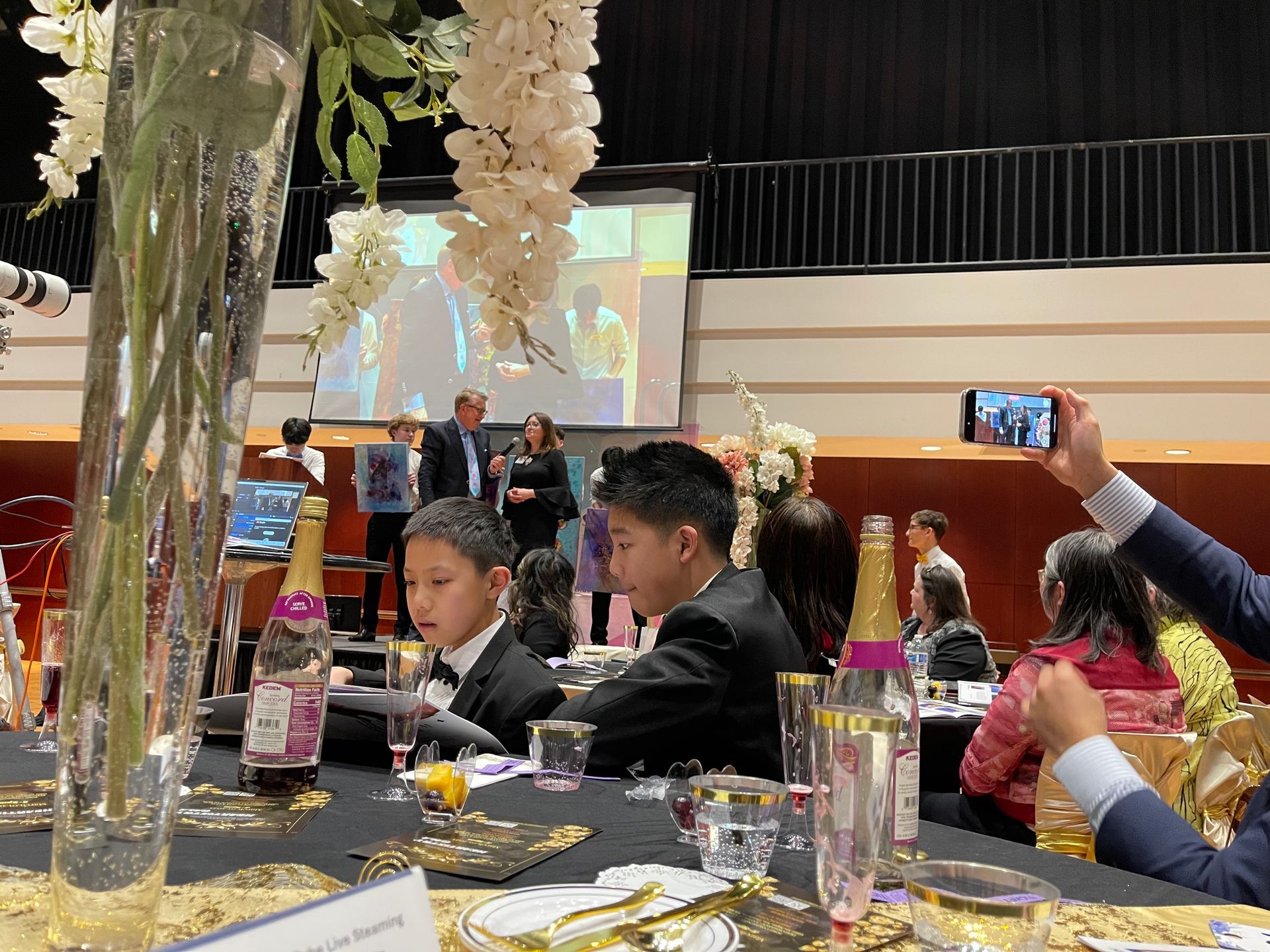 People at a formal event, children in suits at a table, presentation on screen, floral decorations.