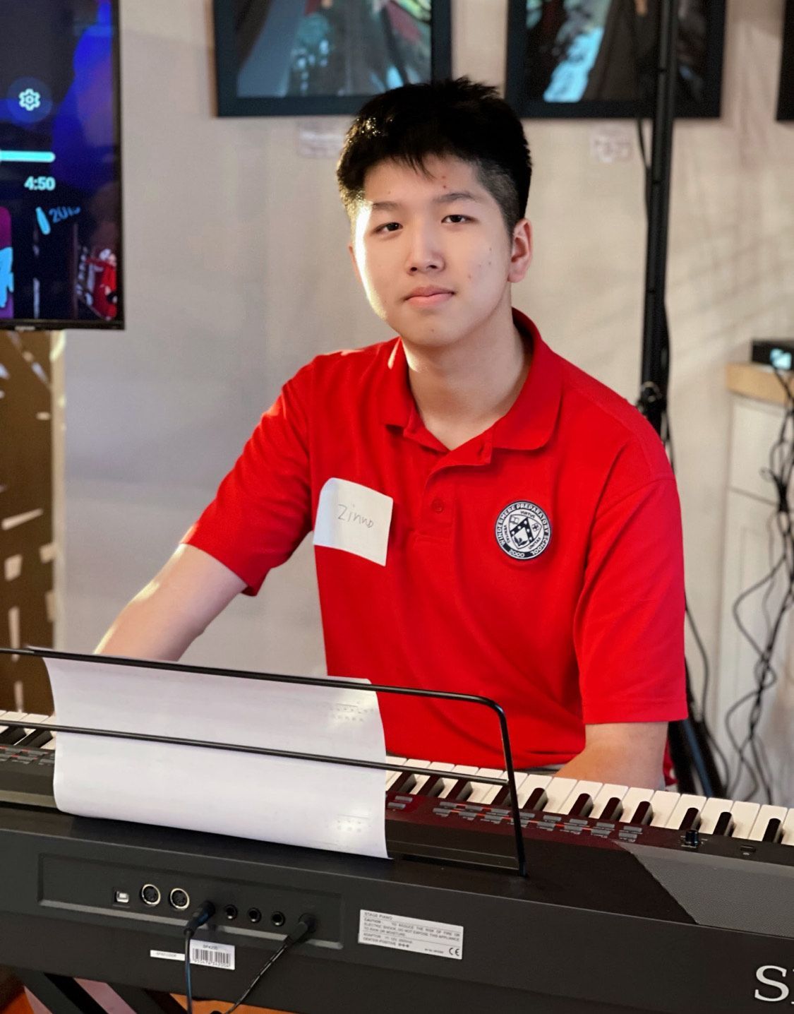 Young man in red shirt at a keyboard, facing camera.  Setting appears to be indoors.