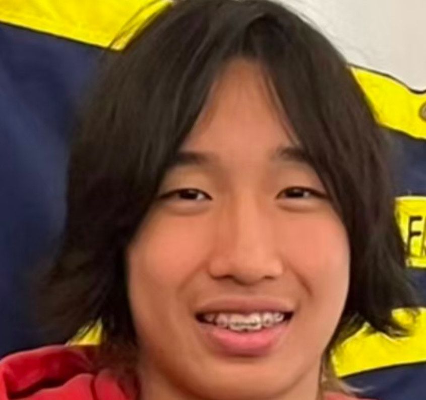 Young Asian person with braces smiles, long dark hair, red hoodie, yellow and blue background.