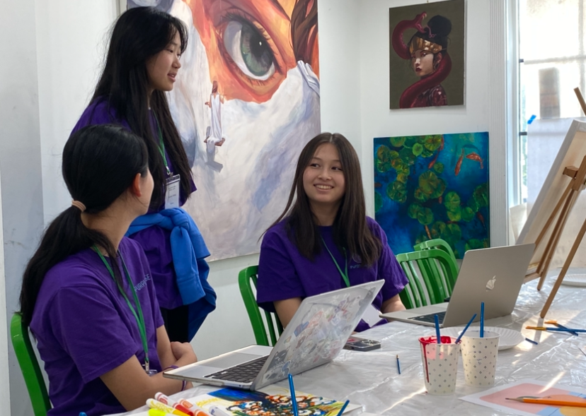 Three young women in purple shirts in art studio, smiling and talking by laptops. Artwork on walls.
