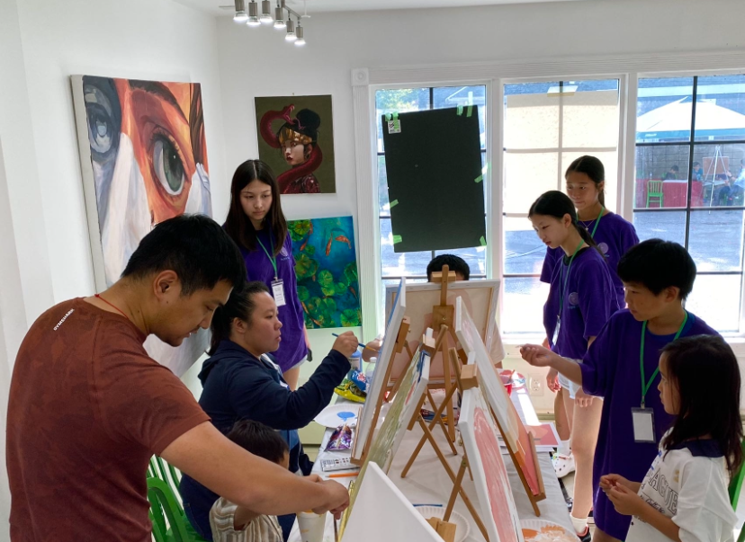 People painting in an art class. Instructor with students, easels with canvases, artwork on walls, and a bright room.