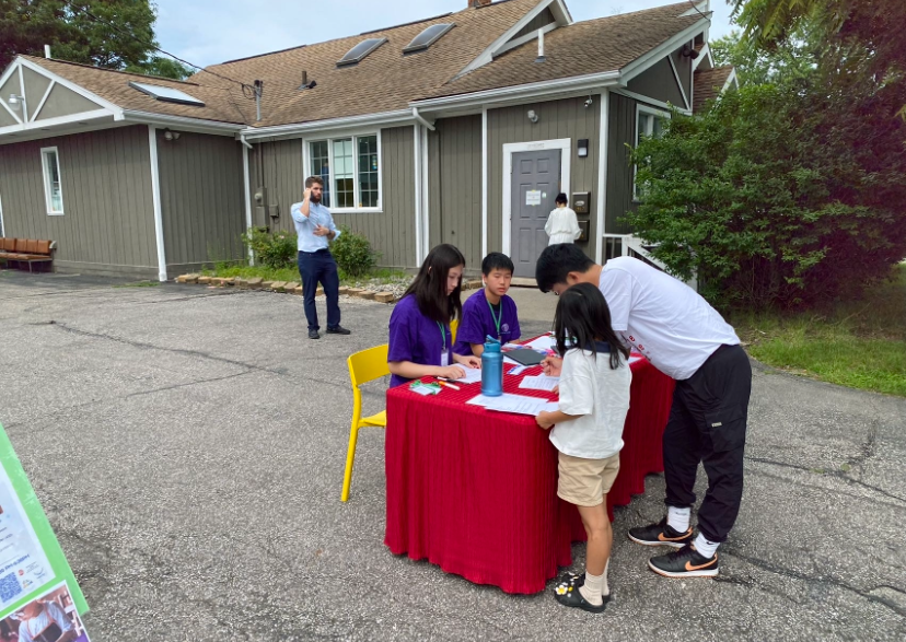 People at an outdoor registration table in front of a building. A person in a suit watches.