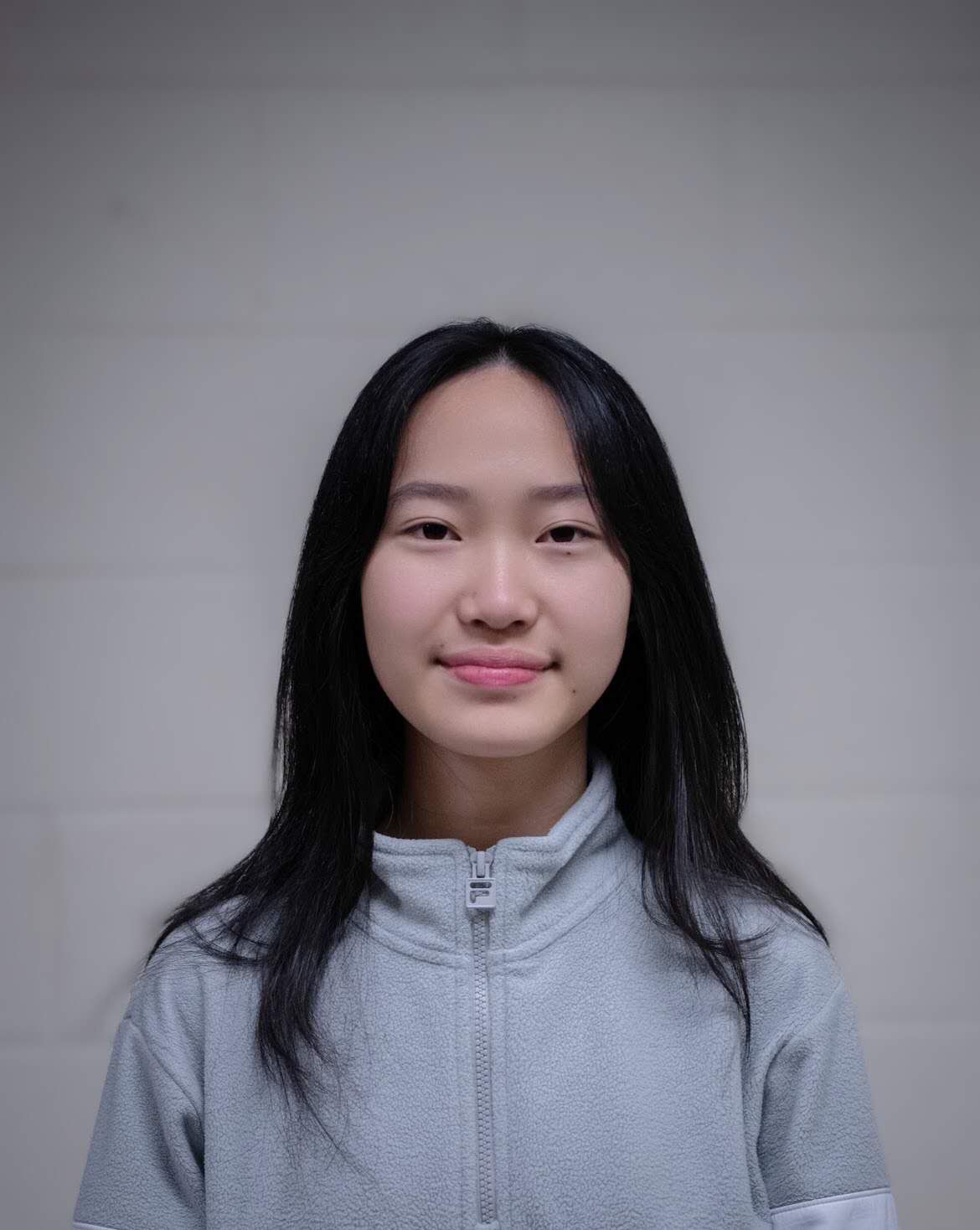 Young Asian woman with long black hair in gray zip-up smiling at the camera in front of a white wall.