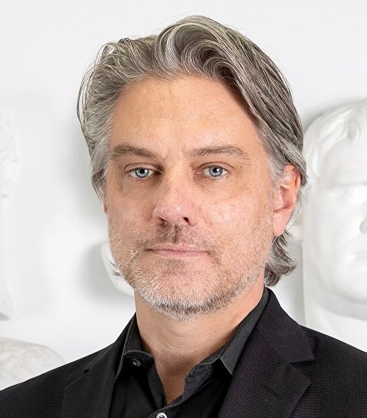 Man with graying hair and beard, wearing a black shirt and jacket, looking at the camera.