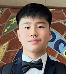 Young man with black hair, bow tie, and suit jacket smiles against a brick background.