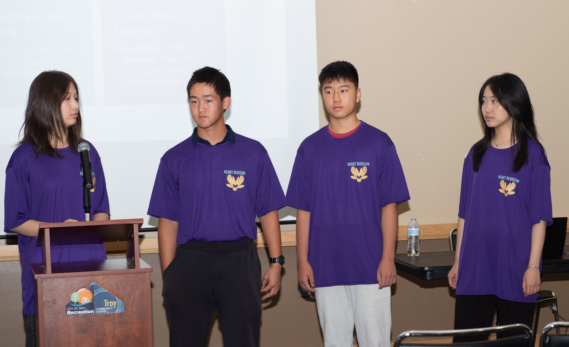 Four students in purple shirts with a logo on stage giving a presentation.