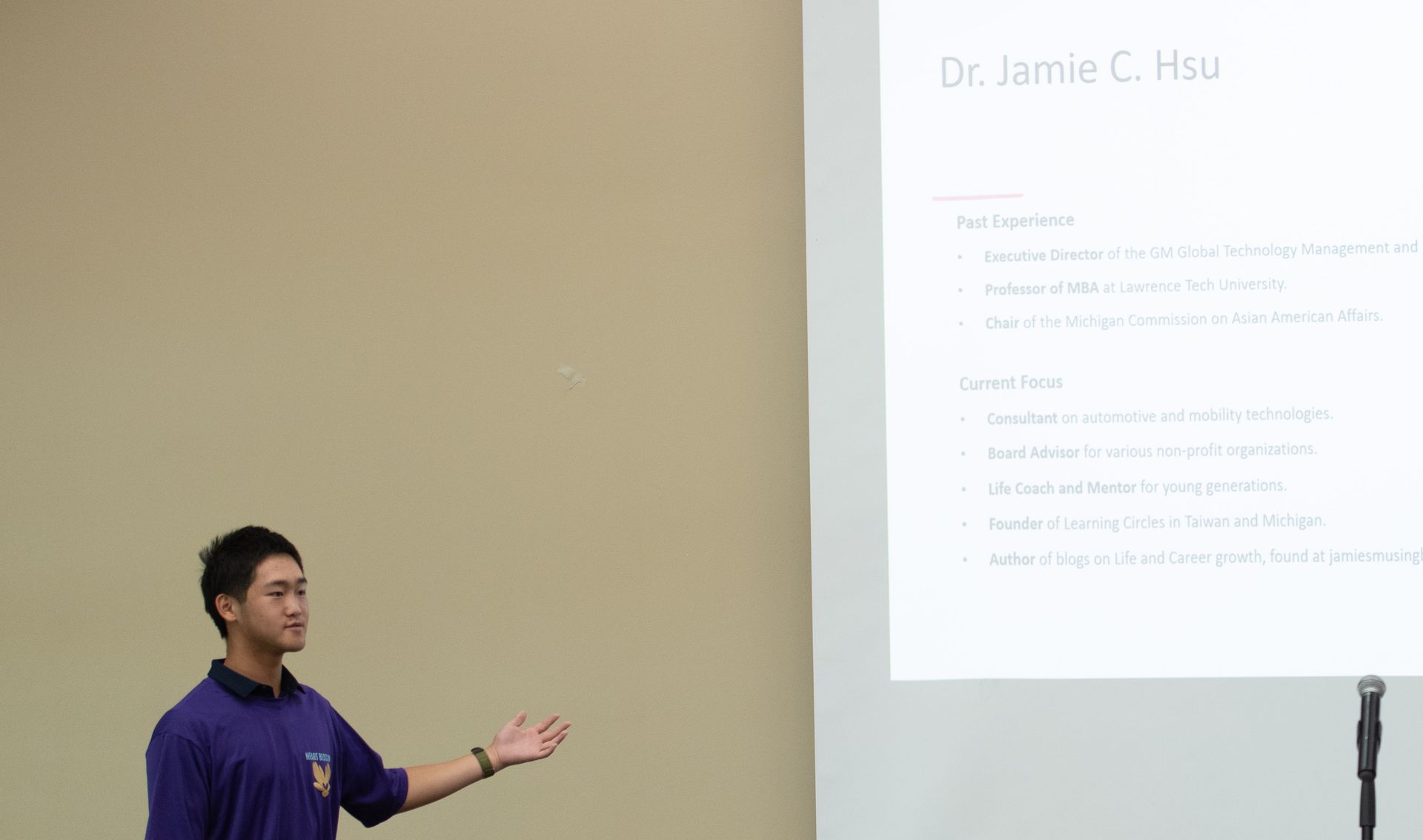 Man in purple shirt gestures toward presentation on screen; cream-colored wall backdrop.