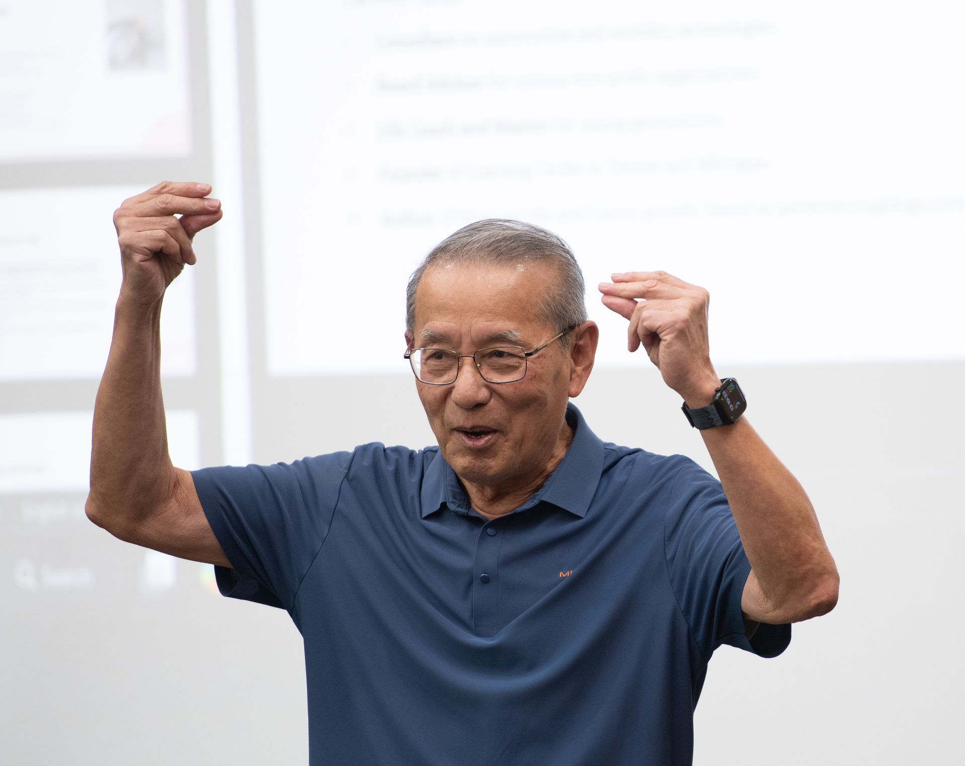 Older man with glasses and a blue shirt gestures with both hands, speaking.