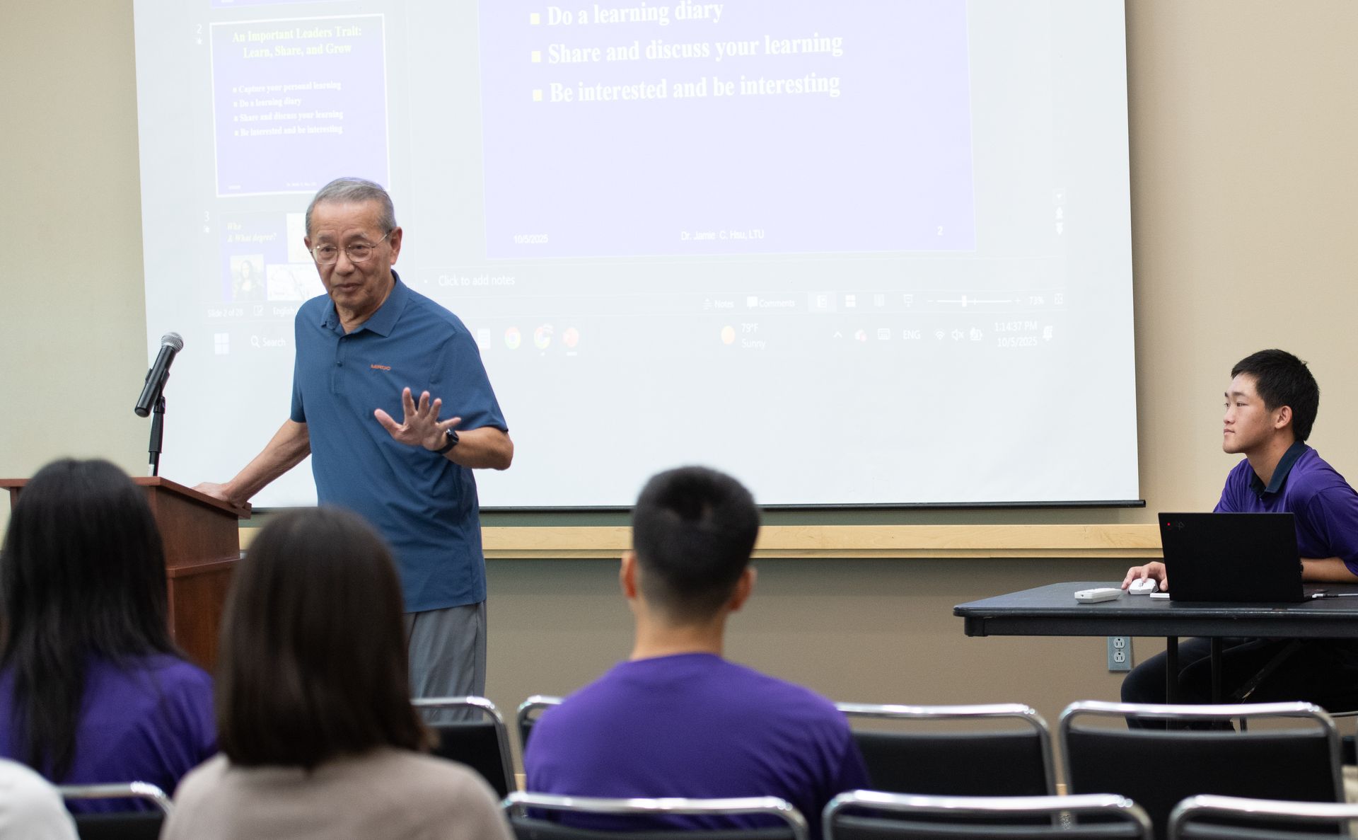 Professor lecturing; students listen in a classroom. Man at a podium gestures, screen behind him.