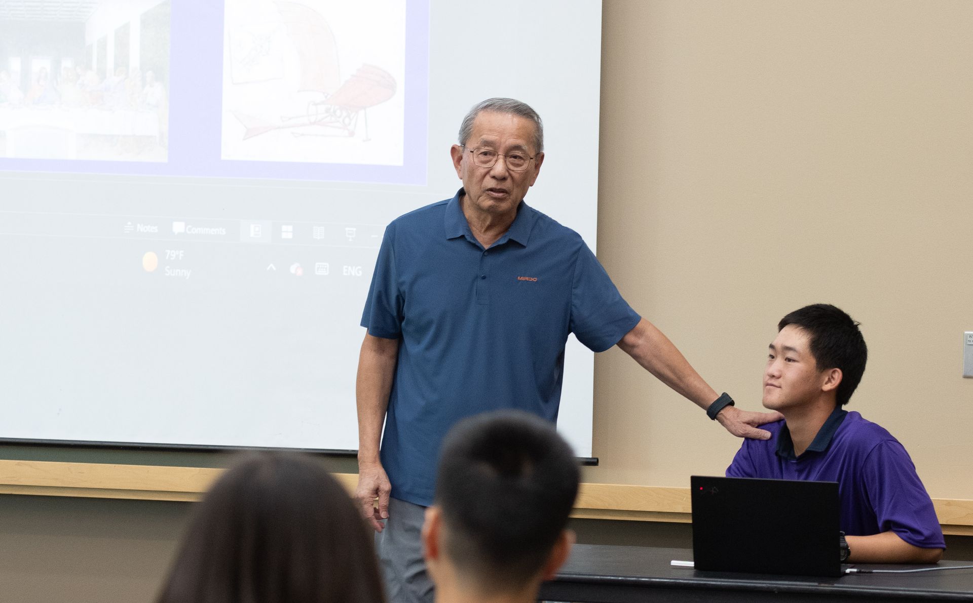 Professor in blue shirt points at student at desk in a classroom.