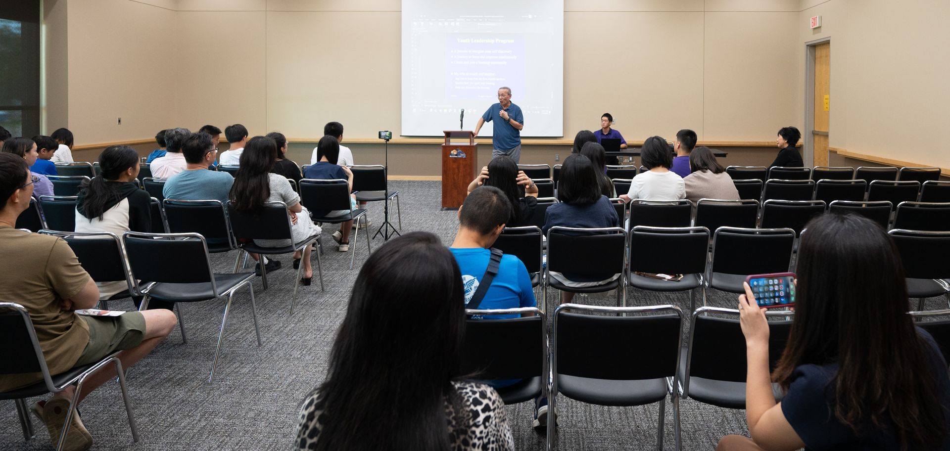 A lecture in progress. A man speaks at a podium to a seated audience in a large room.