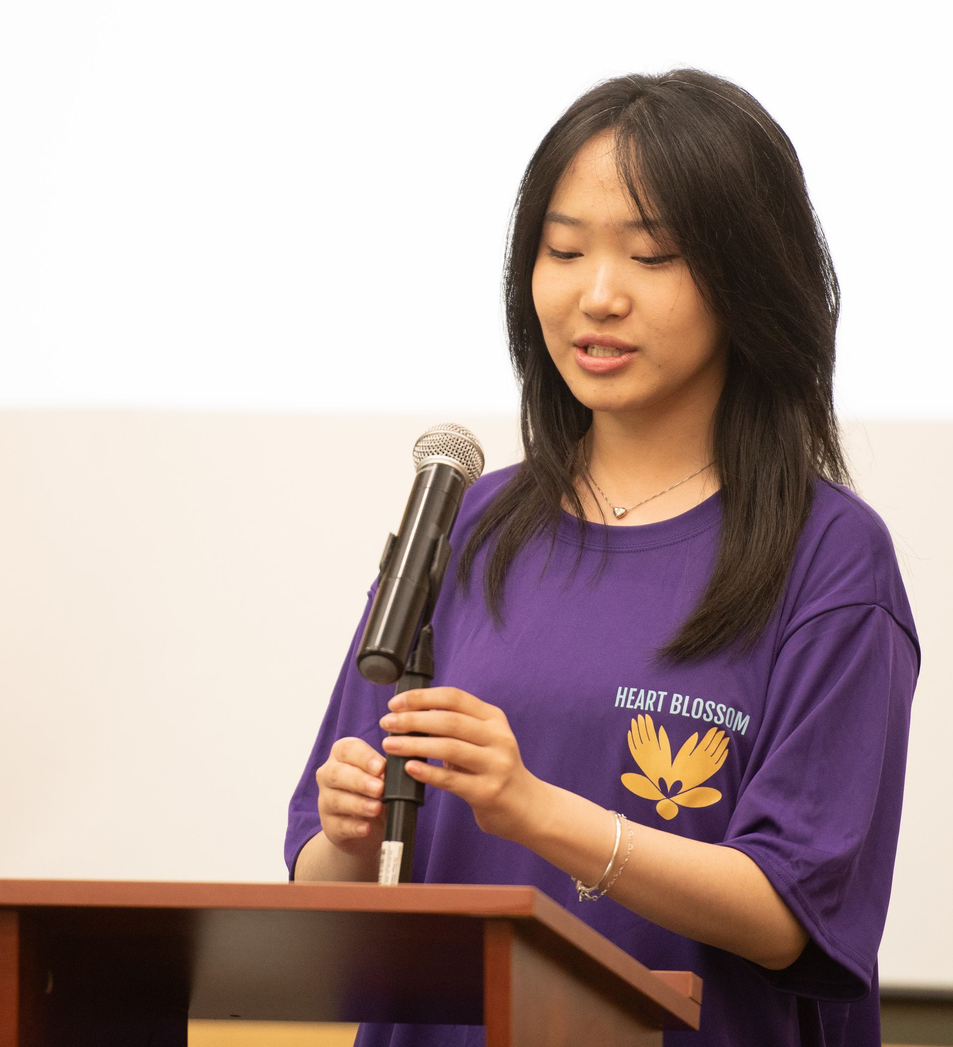 Woman in purple shirt speaking at a podium with a microphone. Light-colored background.