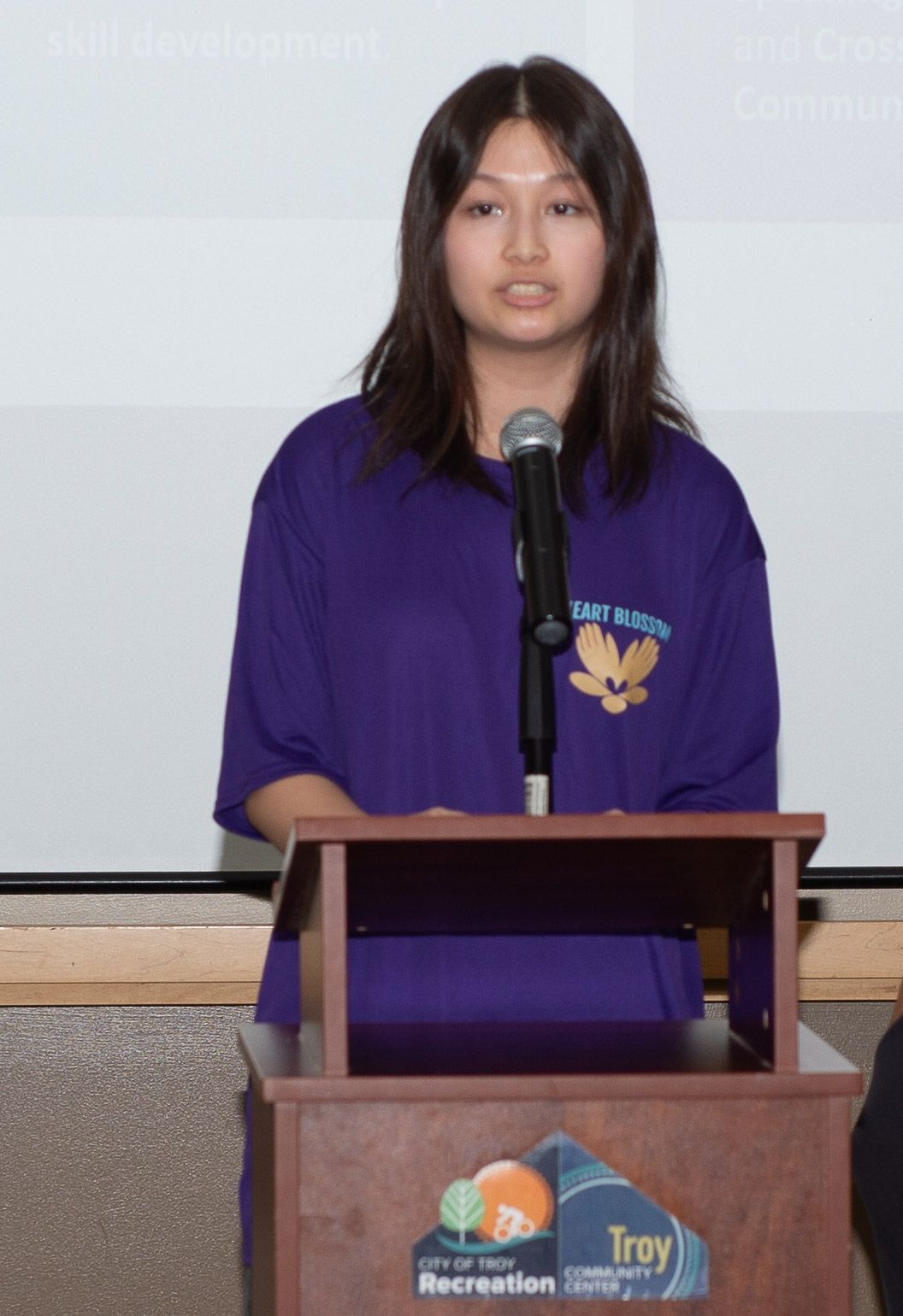 Woman in purple shirt speaks at podium, Troy Recreation logo visible.