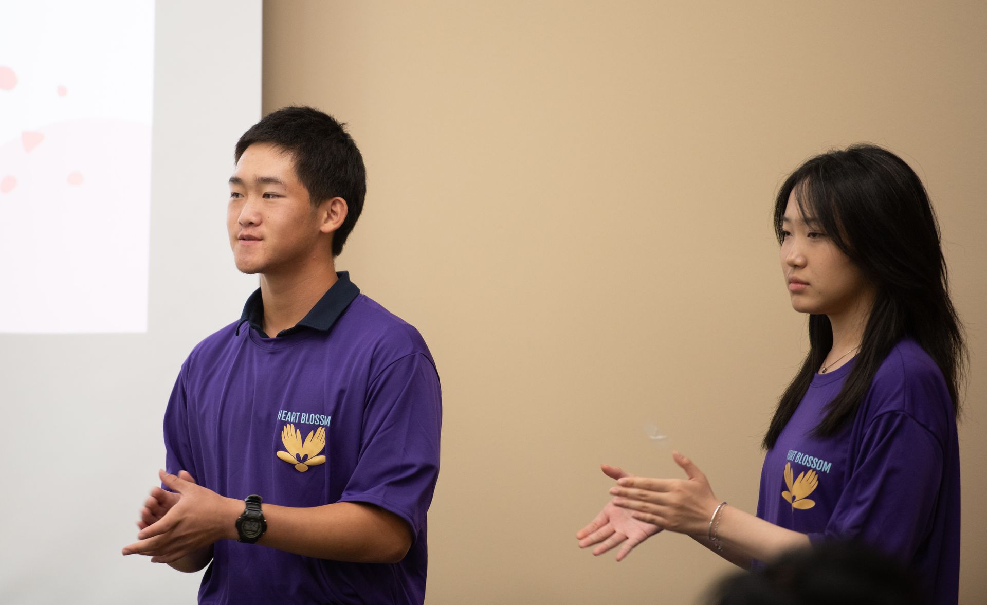 Two young people in purple shirts clapping during a presentation. Light-colored wall background.