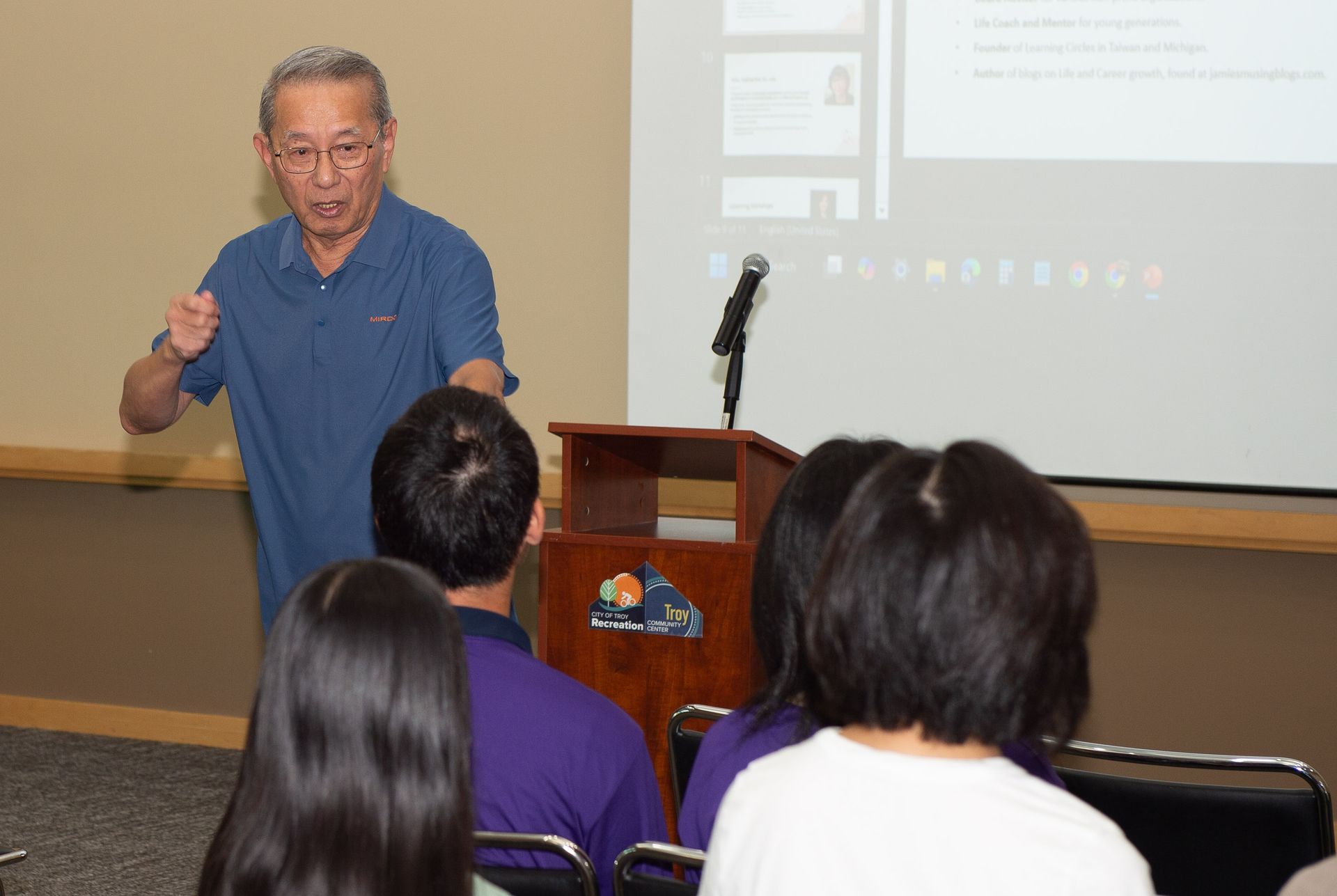 Man in blue shirt giving a presentation to a small audience in a beige room, gesturing with his hand.
