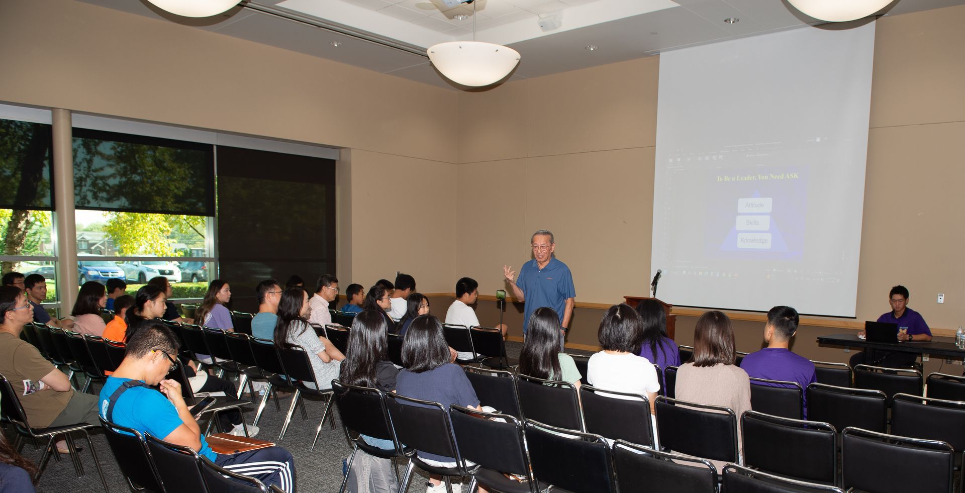 A man presenting to a group of people in a lecture hall with a projector screen.