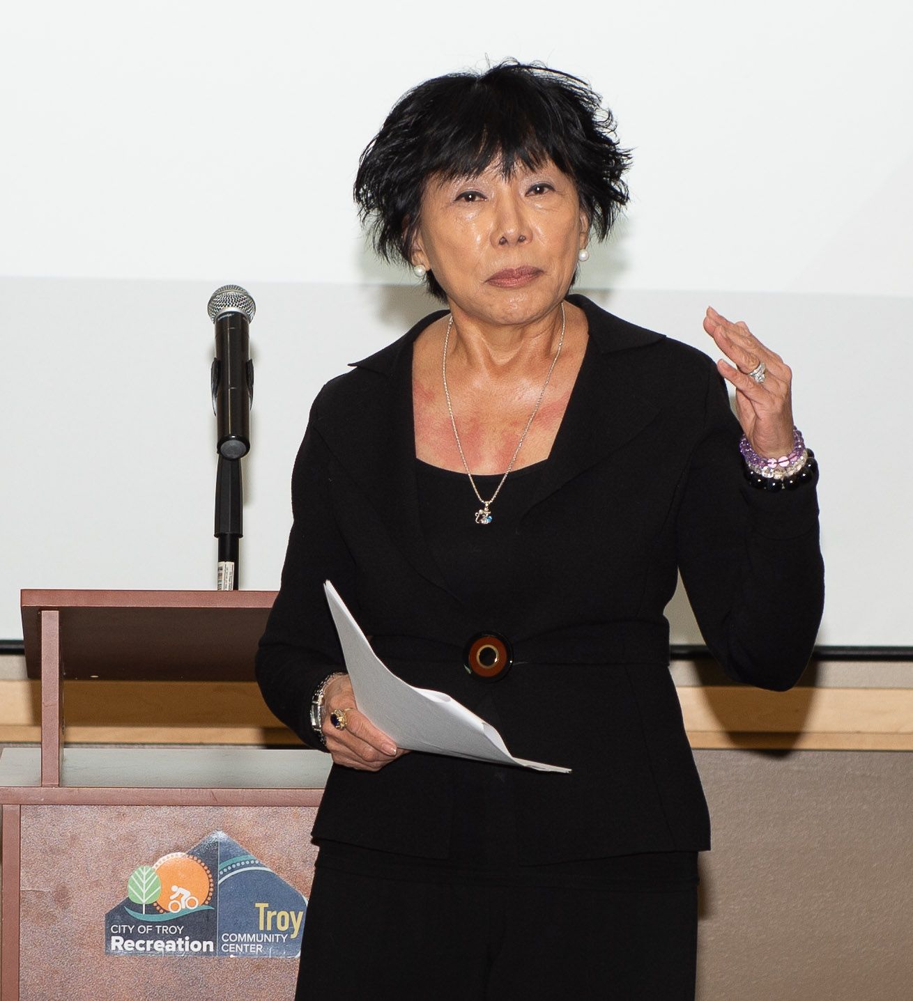 Woman with dark hair speaking at a podium, holding papers, gesturing, wearing a black outfit, standing indoors.