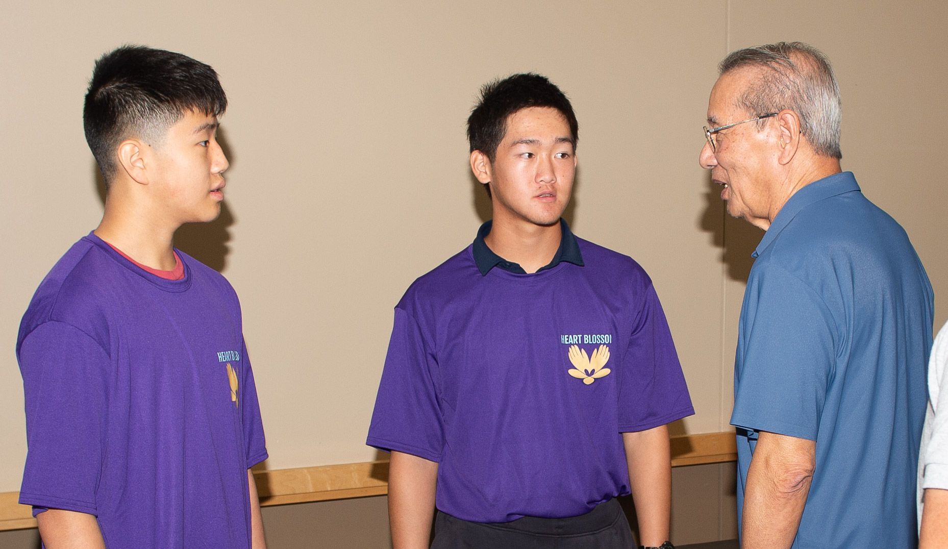 Two young men in purple shirts converse with an older man in a blue shirt in a room with a beige wall.
