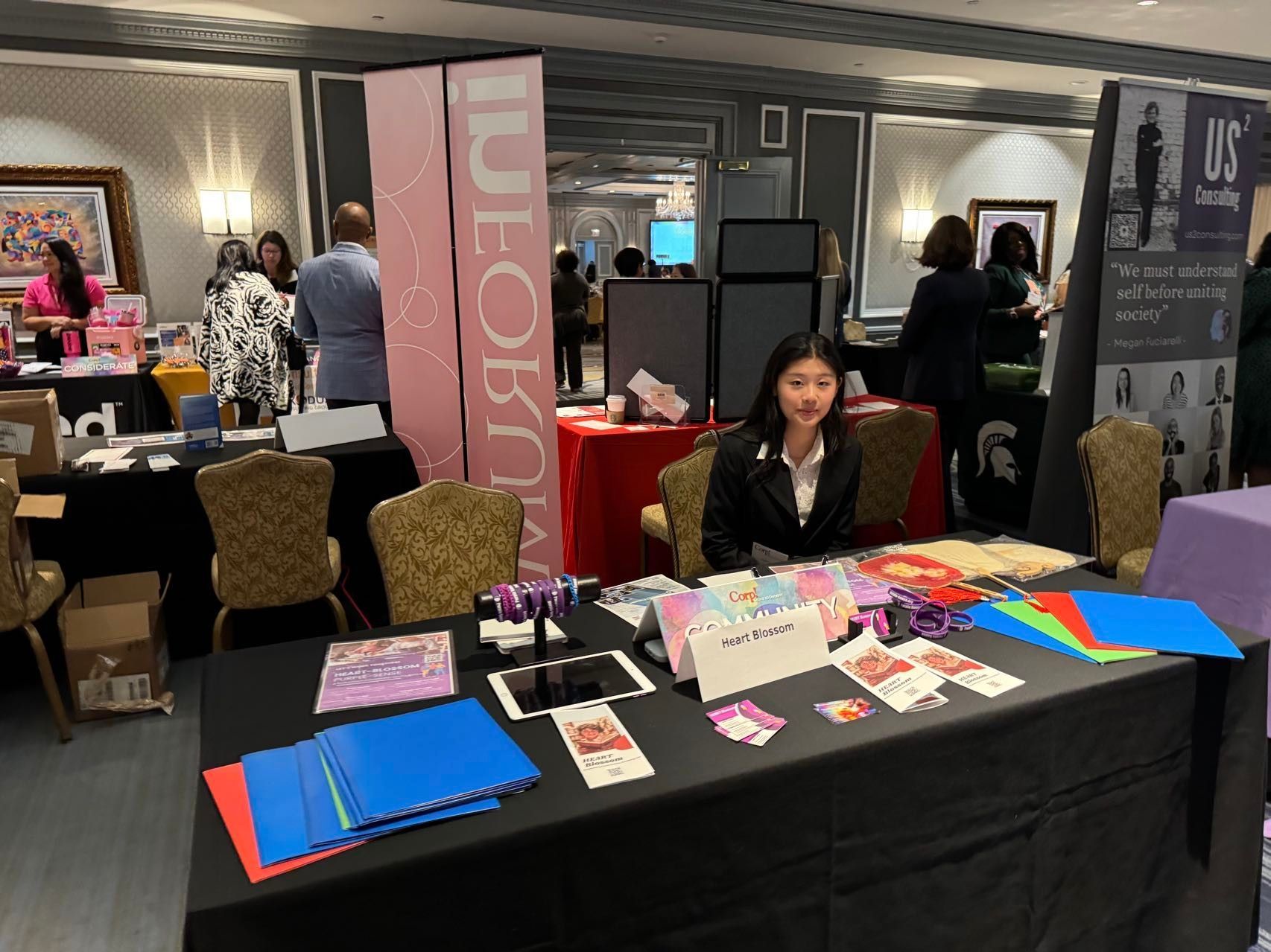 A woman sits at a vendor table with products displayed. Event setting, featuring signs and displays.