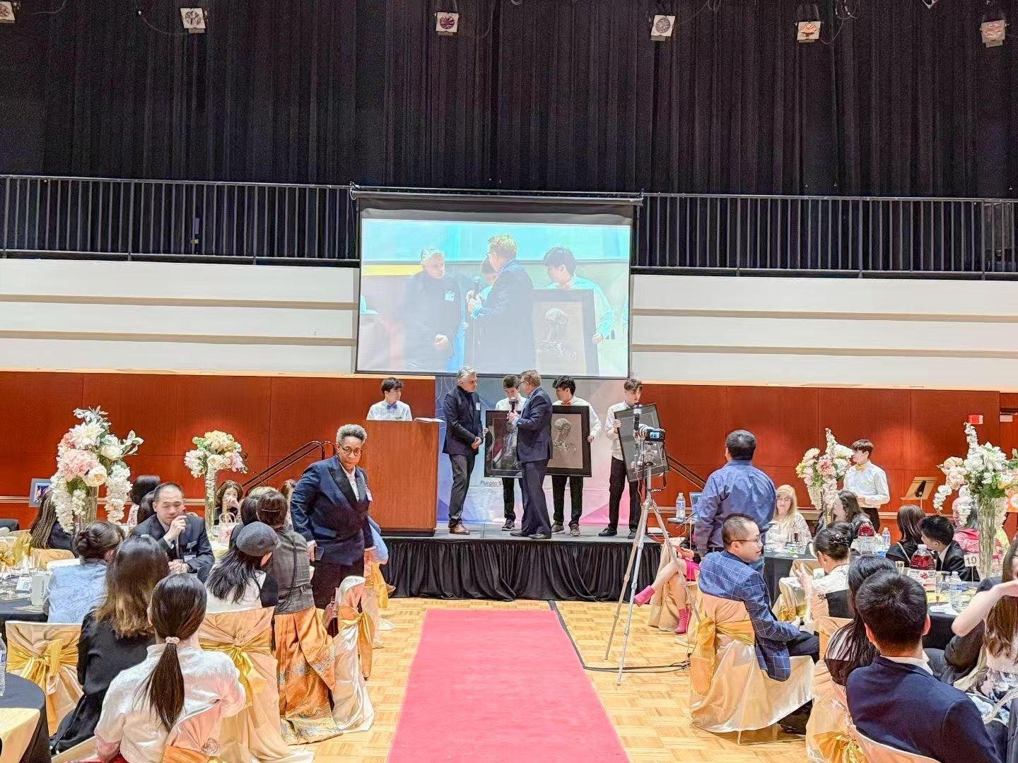 People on stage at an event, presentation of an award, audience in the background, red carpet.