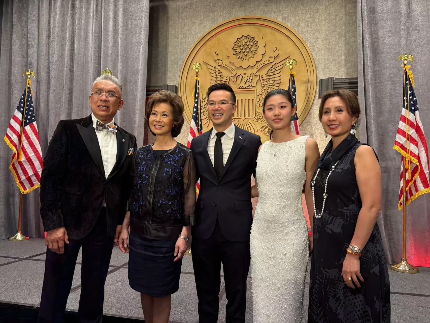 Group of five people posing at an event with US flags and seal backdrop. Formal attire.