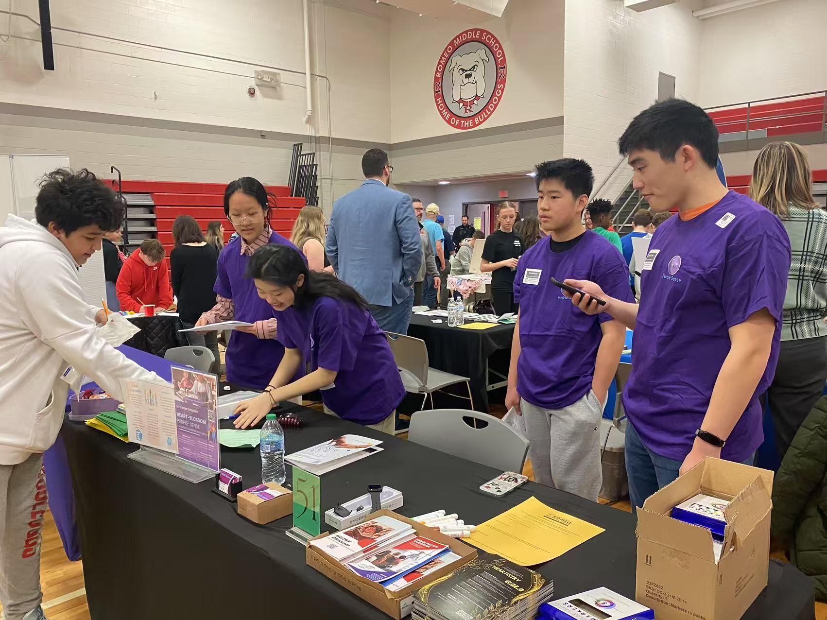Students at a table, setting up for an event, wearing purple shirts. Gym setting with red trim.