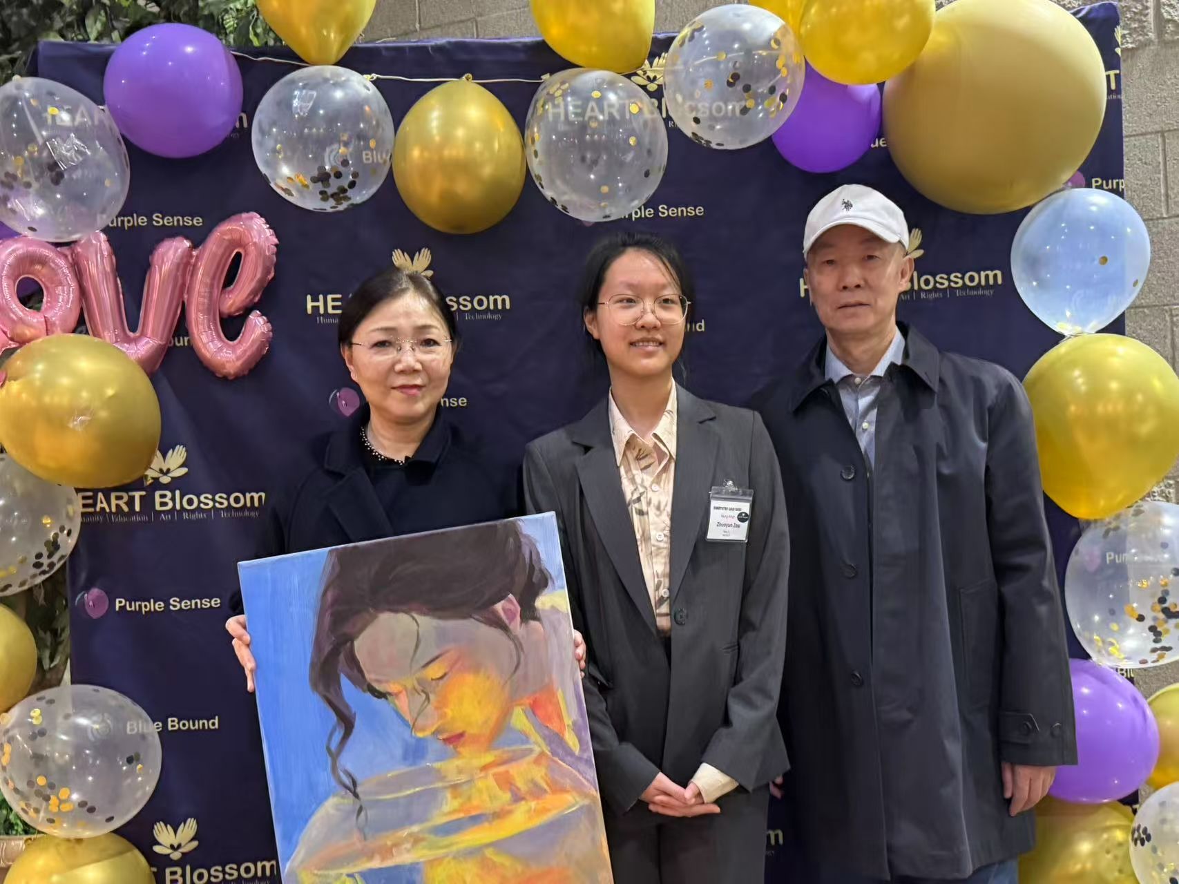 Three people pose, holding a painting, in front of balloons. Woman, teen, man stand at event with “love” backdrop.