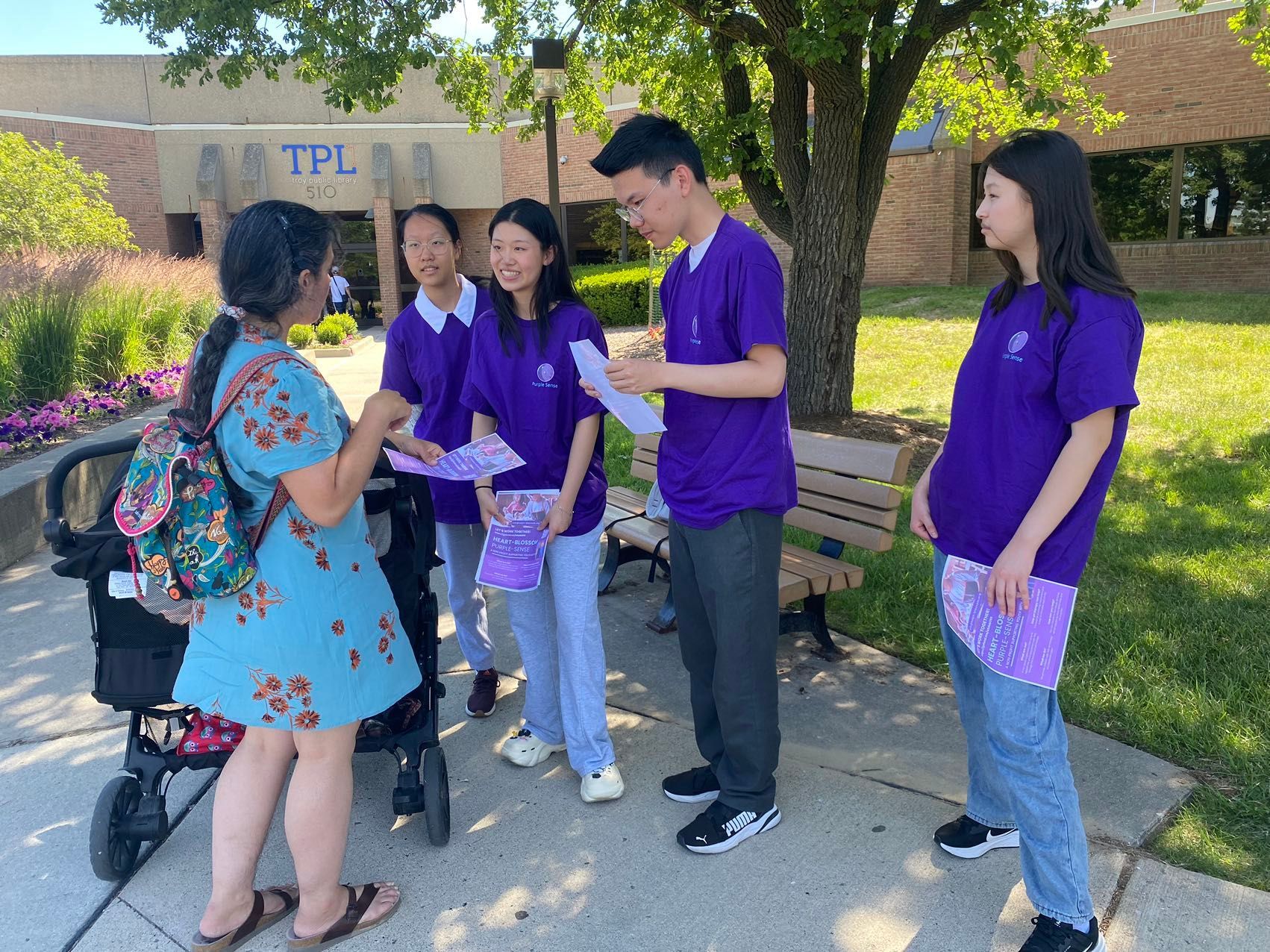 Five people in purple shirts handing out flyers outside of a building, speaking to a woman with a stroller.