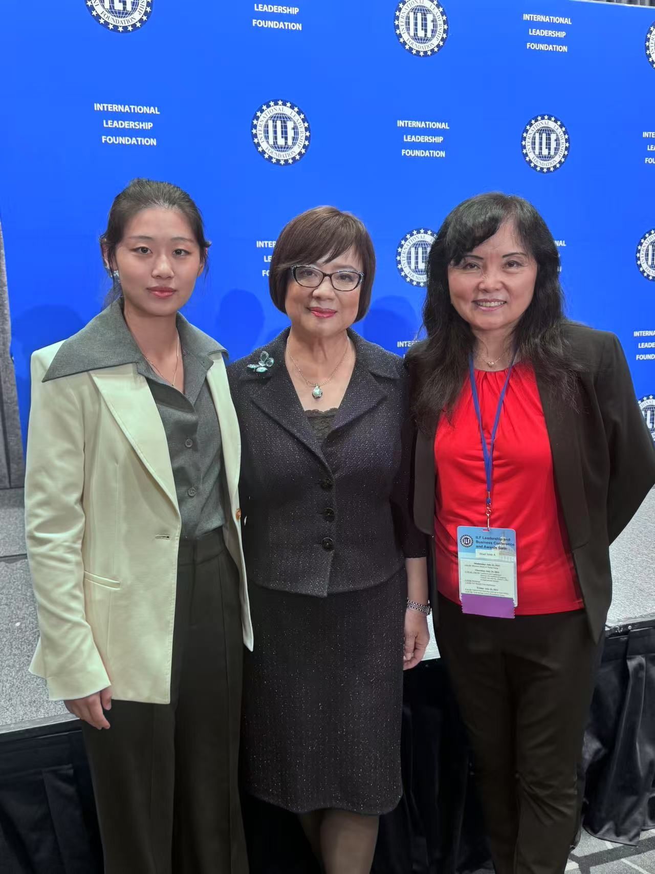 Three Asian women pose together; blue backdrop, dressed professionally, smiling.