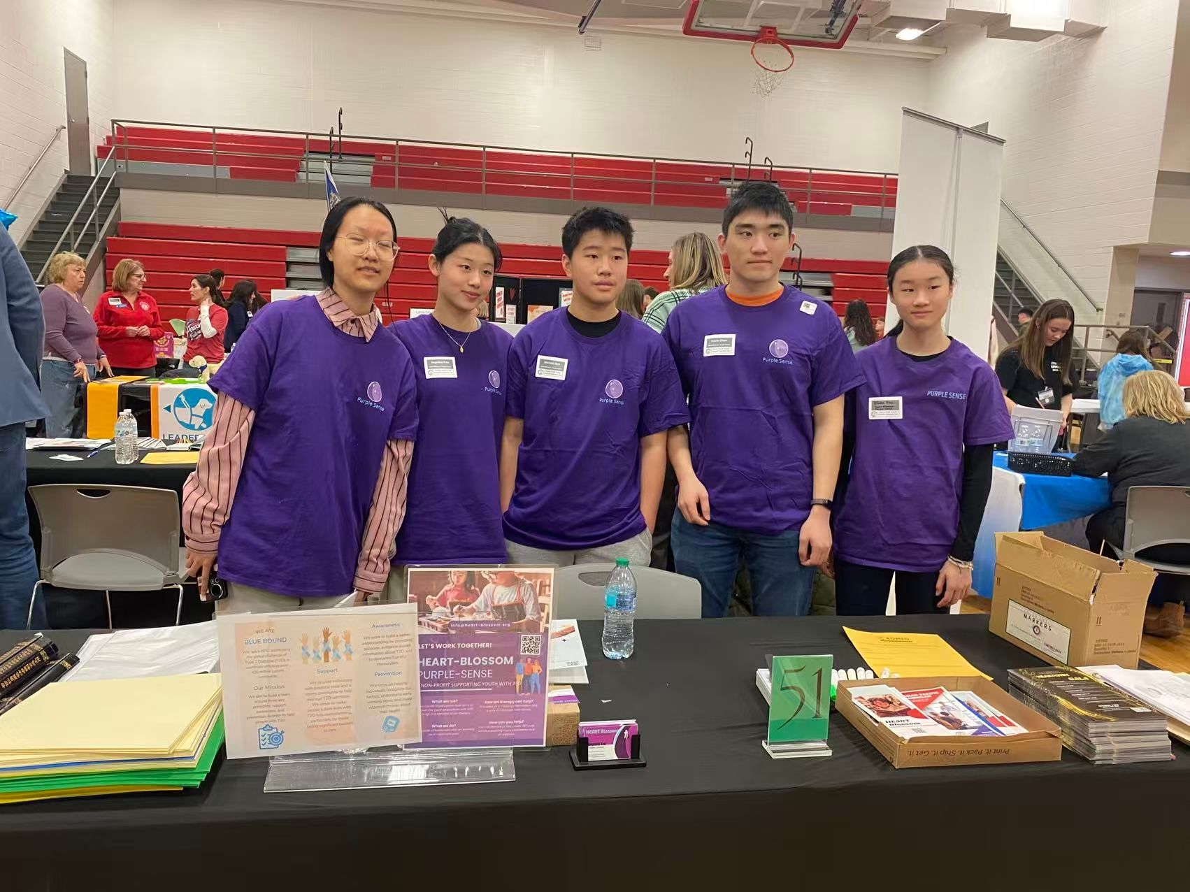 Five people in purple shirts stand behind a table, likely at an event.