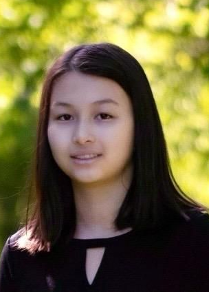 Young woman with dark hair in front of a blurred green tree backdrop. She wears a black shirt and smiles slightly.