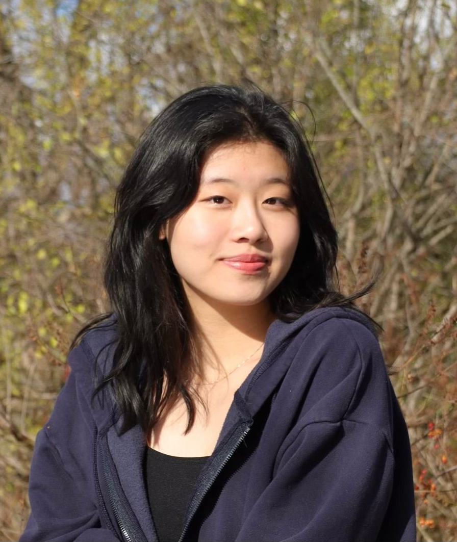 Young Asian woman in a blue hoodie smiles outside, trees in the background.