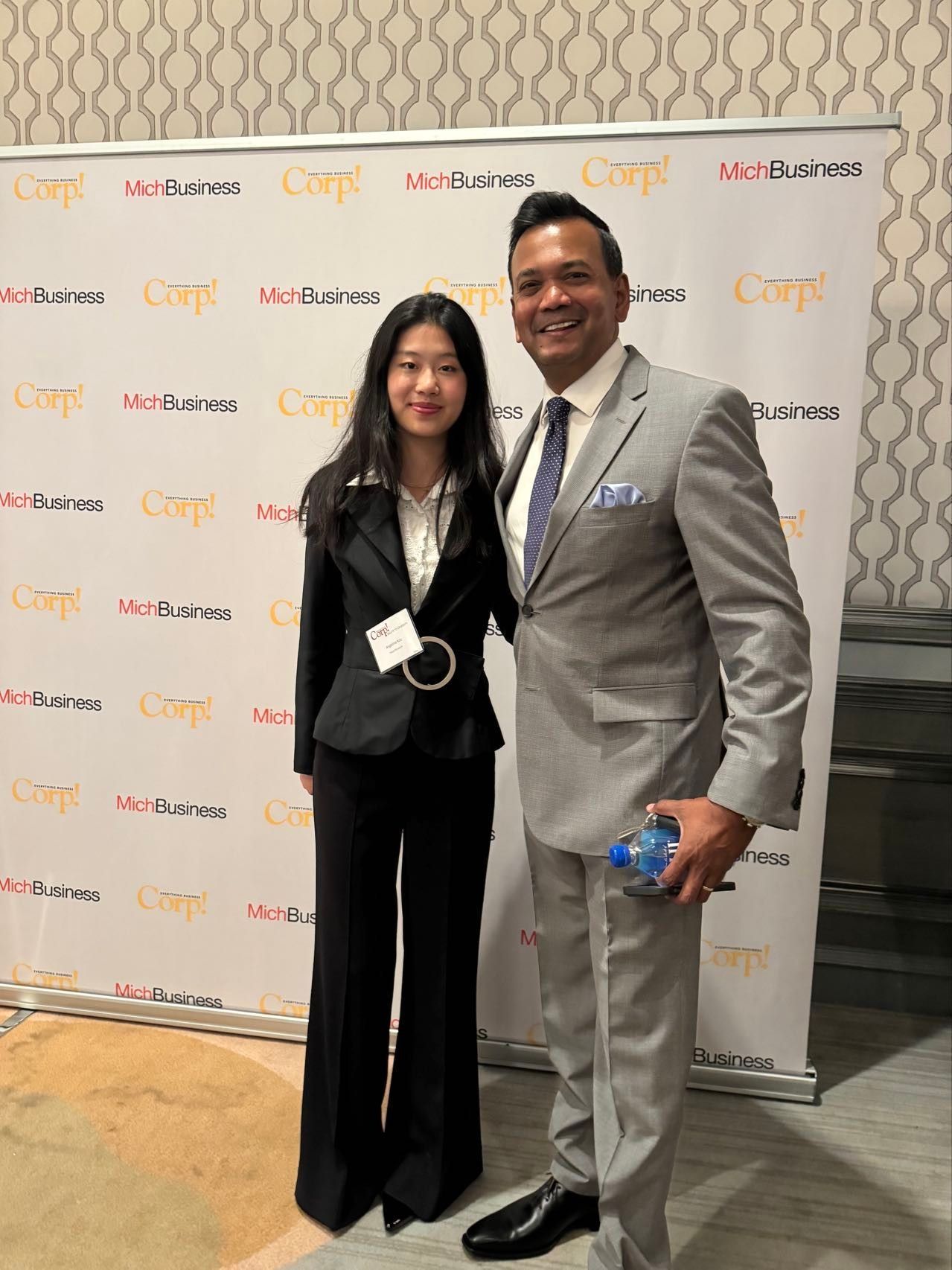 Woman and man pose in front of a backdrop with logos; woman in black suit, man in grey suit.
