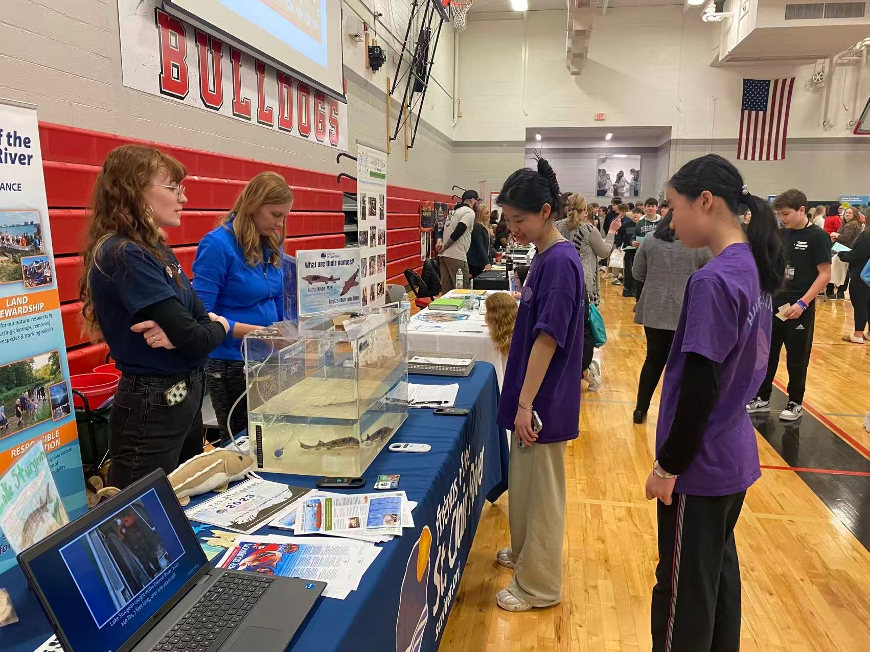 People at a booth with a fish tank in a gymnasium. Two women and two girls are looking at the display.