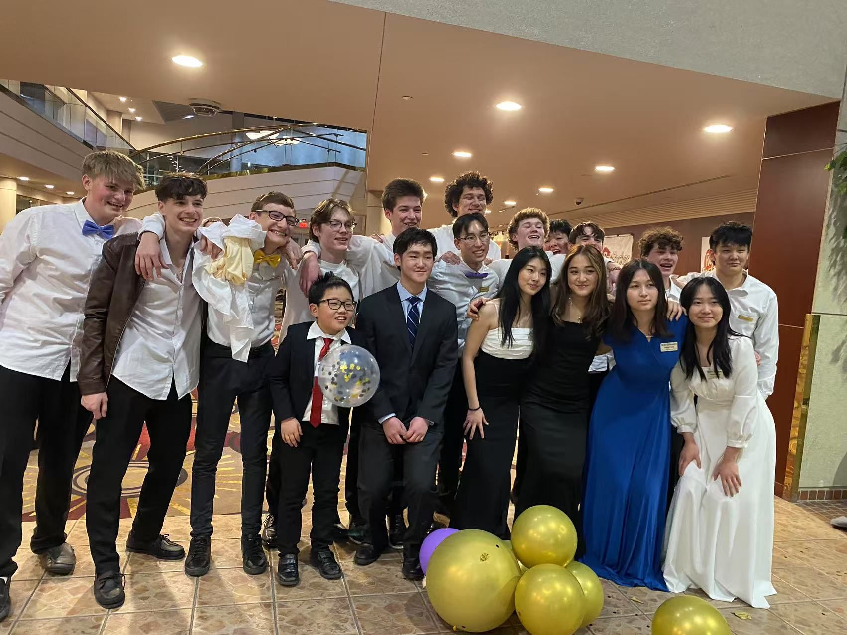 Group of teenagers in formal wear, smiling, posing for a photo in a building lobby with balloons.