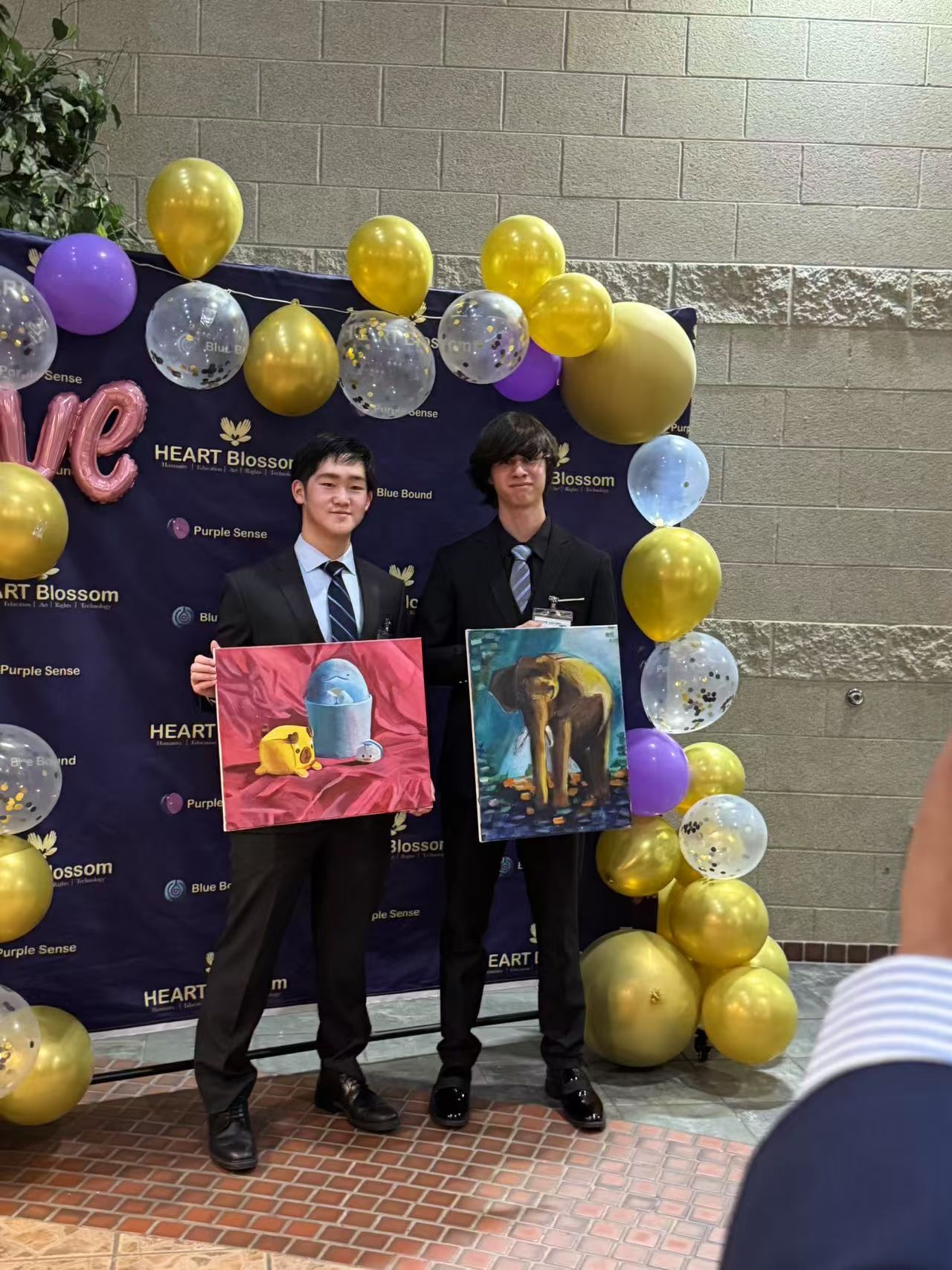 Two young men in suits hold art at an event with balloons. One is smiling. Gold and purple decor.