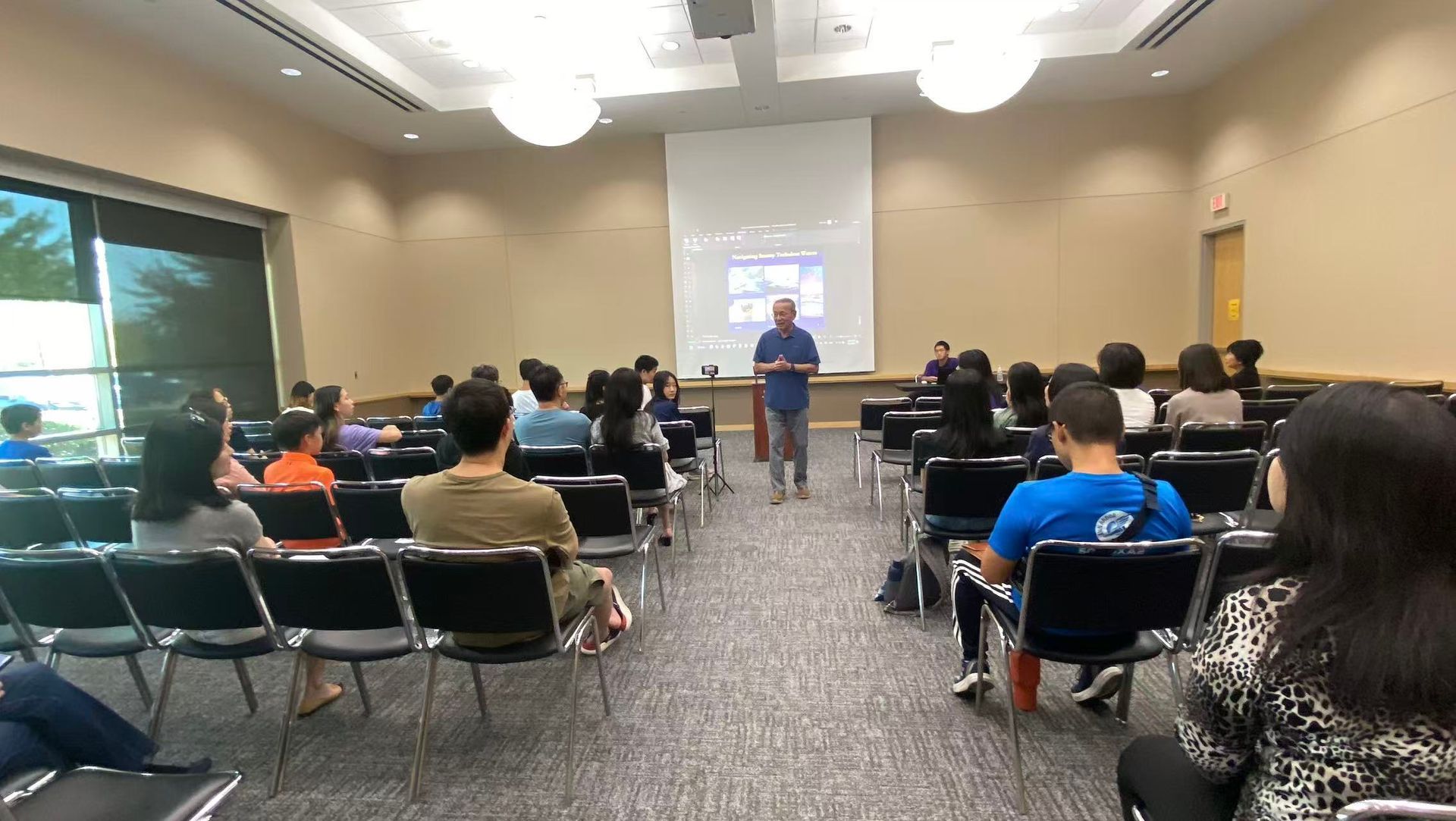 A speaker addresses a group of people seated in a large room during a presentation.