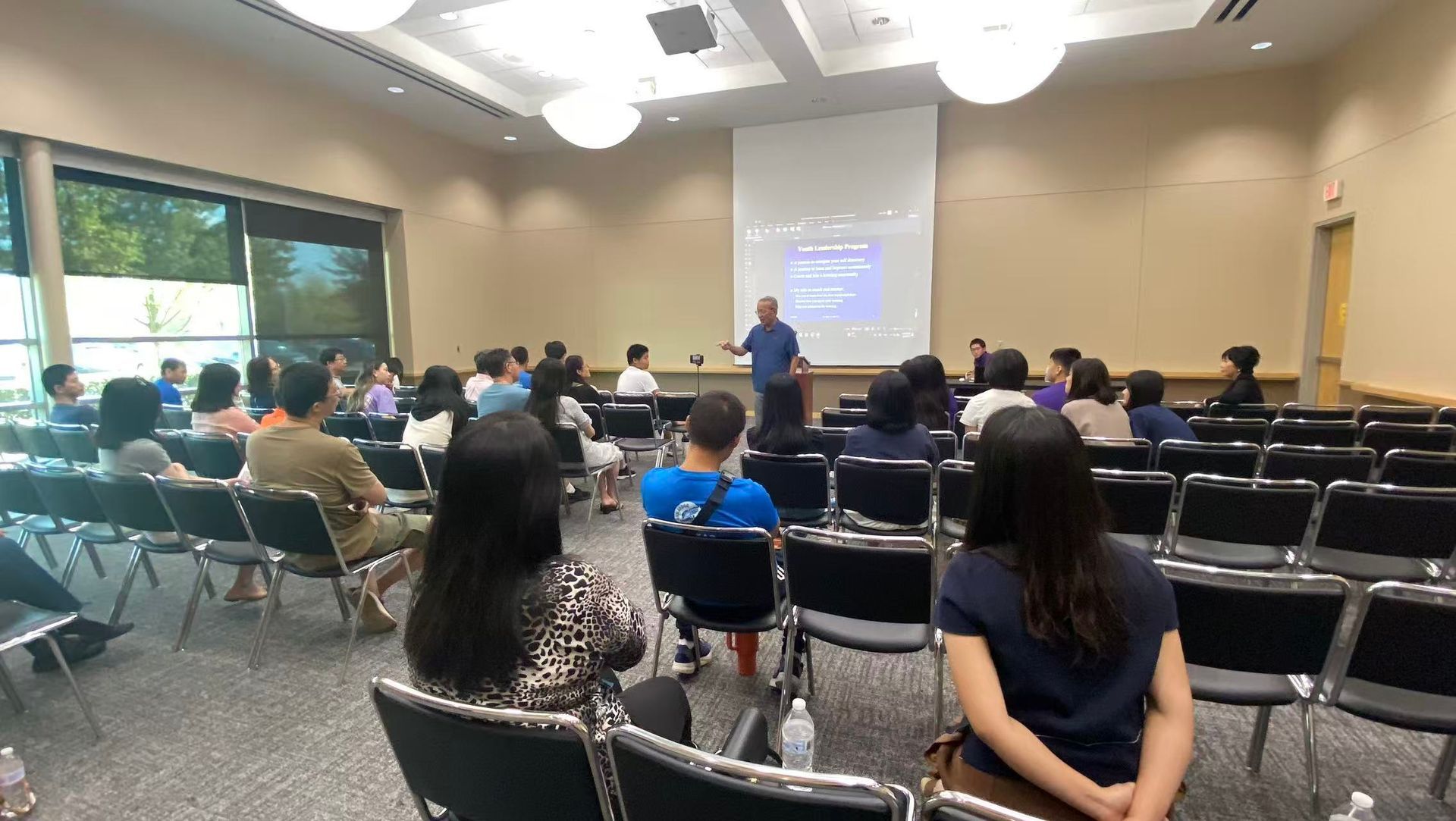 A presenter stands before a projected screen addressing a crowd in a large conference room.