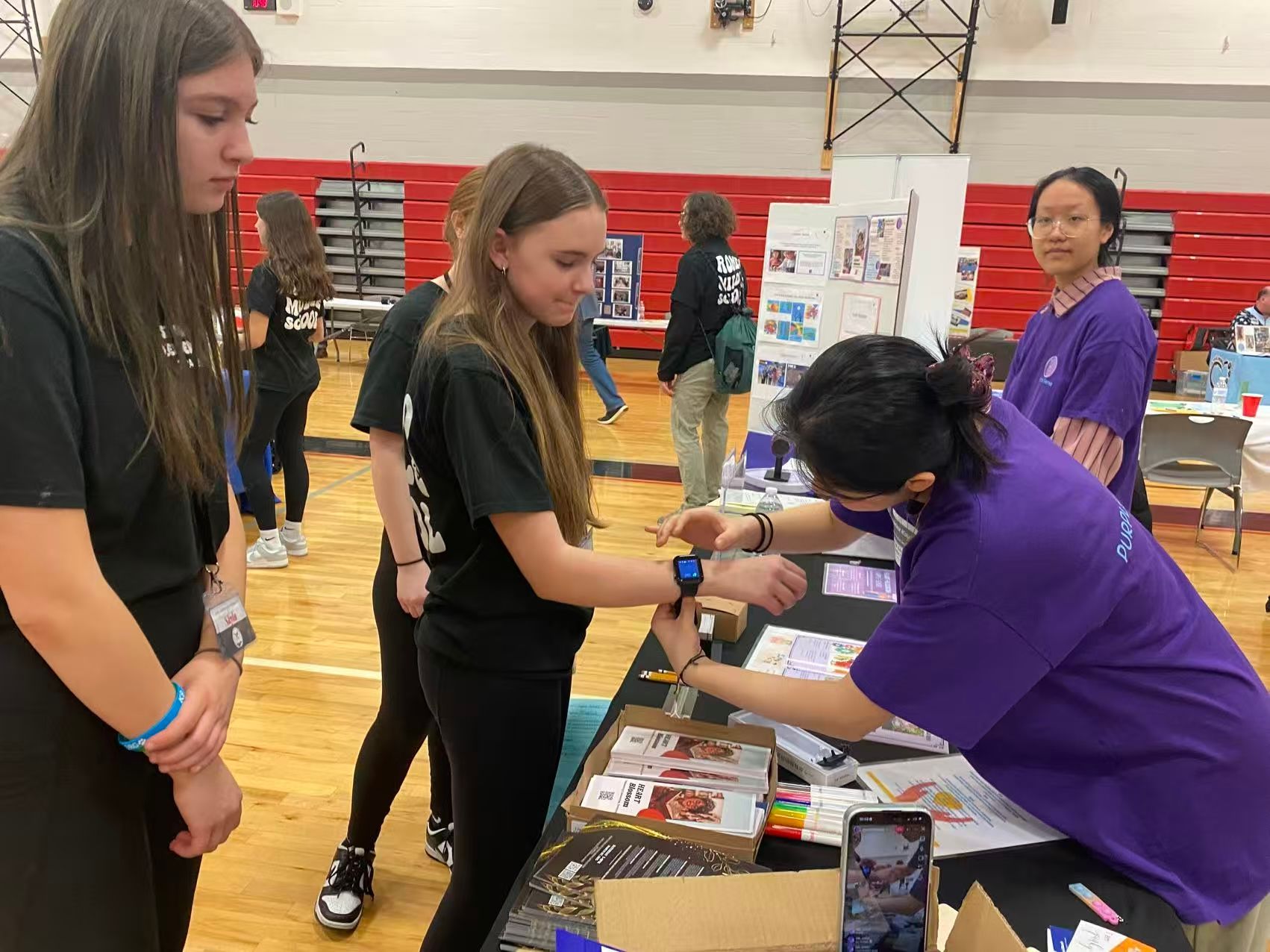 Teenagers at a table, one wears a smartwatch while another looks on. An adult assists at a school event.