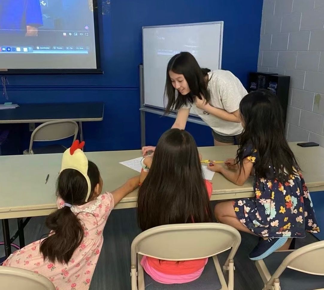Woman assisting three children at a table in a classroom. One child has a chicken hat.