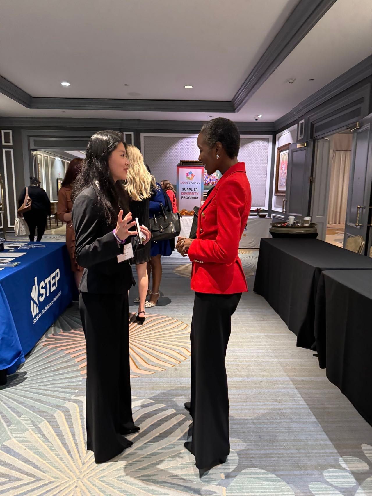 Two women conversing at an event, one in a red blazer, the other in a black suit.