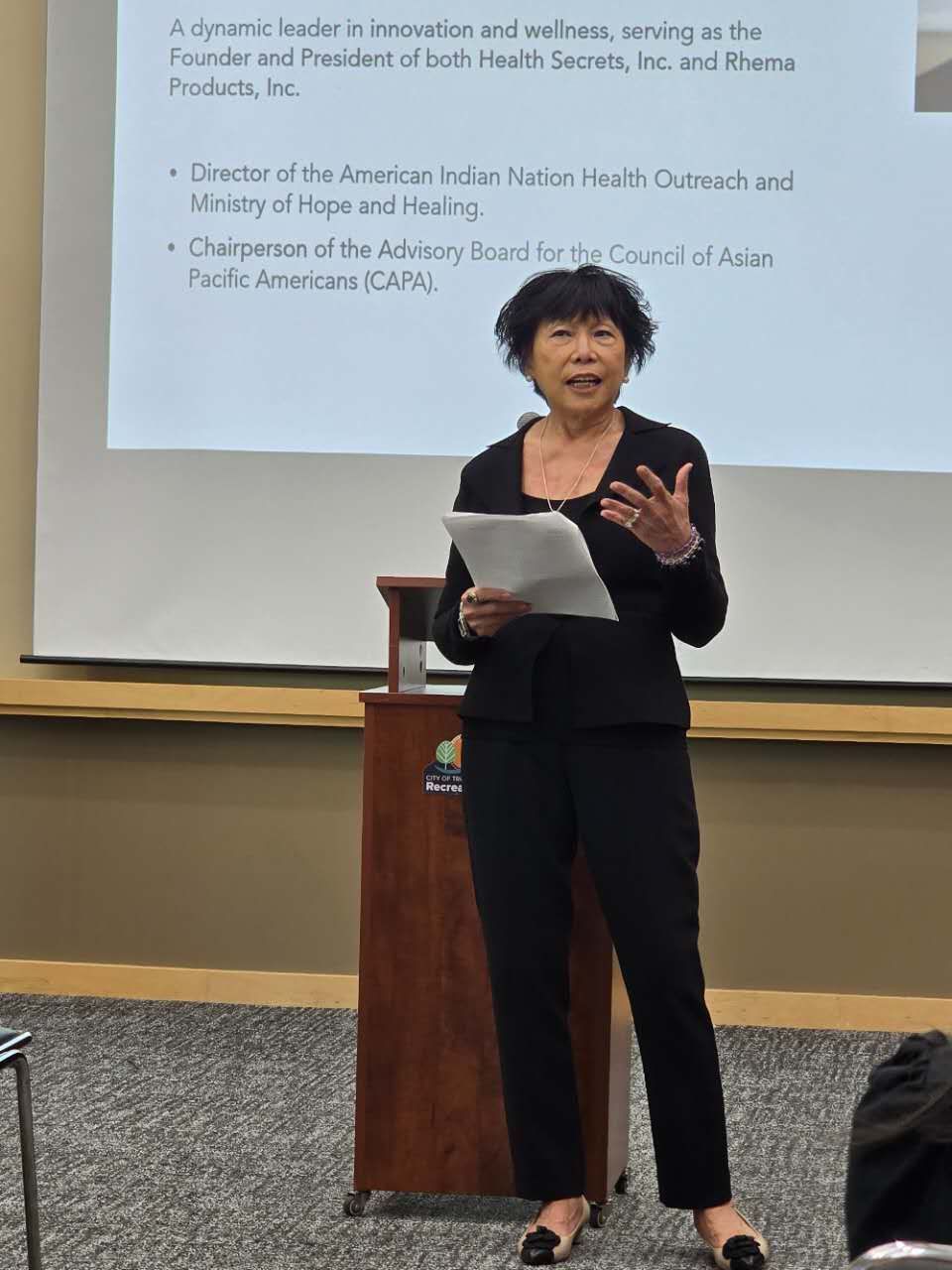 Woman with dark hair giving a presentation, standing at a podium in a room, holding papers.