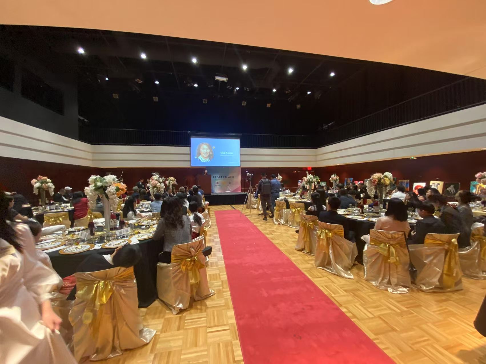 Red carpet event, tables set for dinner, people seated, stage with screen in background.